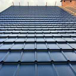 BYX-DACH Przemysław Byczyński - Częściowo pokryty, ciemny dach z blachy trapezowej, widoczny komin z cegły i fragment konstrukcji drewnianej. Ujęcie z dołu, podkreślające regularność wzoru.