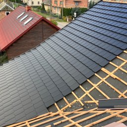 BYX-DACH Przemysław Byczyński - Częściowo pokryty dach blachodachówką w kolorze grafitowym, widoczne łaty i membrana dachowa. W tle budynki mieszkalne i dach z czerwonej dachówki.