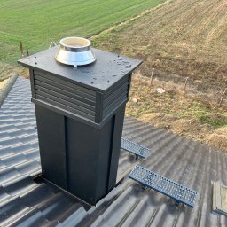 BYX-DACH Przemysław Byczyński - Czarny komin z metalowym zakończeniem na błyszczącym, ciemnym dachu z dachówkami. W tle pole i ogrodzenie. Widoczne stopnie kominiarskie.