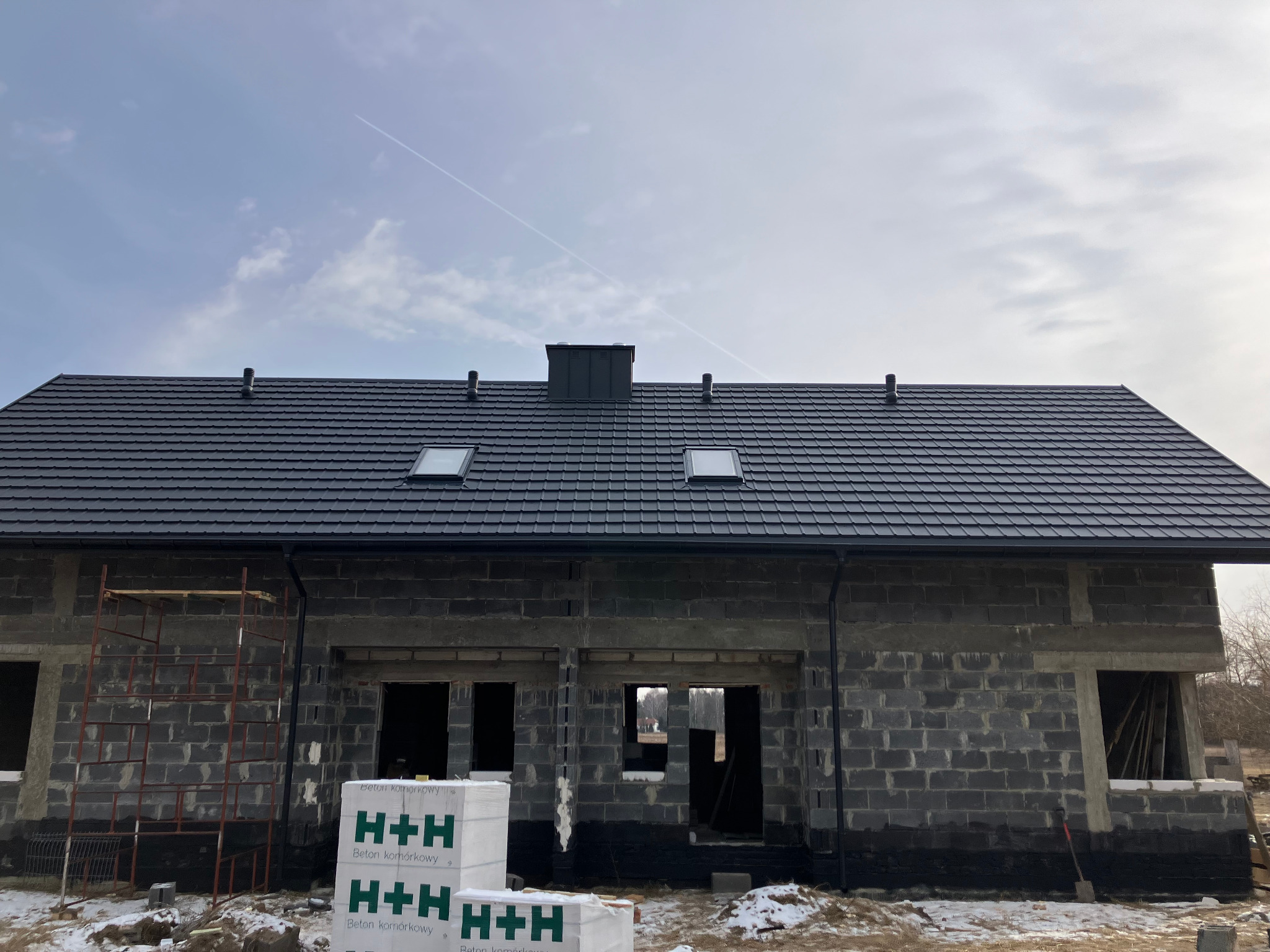 Dom w trakcie budowy z nowym, grafitowym dachem z oknami dachowymi i kominem. Na pierwszym planie palety z betonem komórkowym H+H.