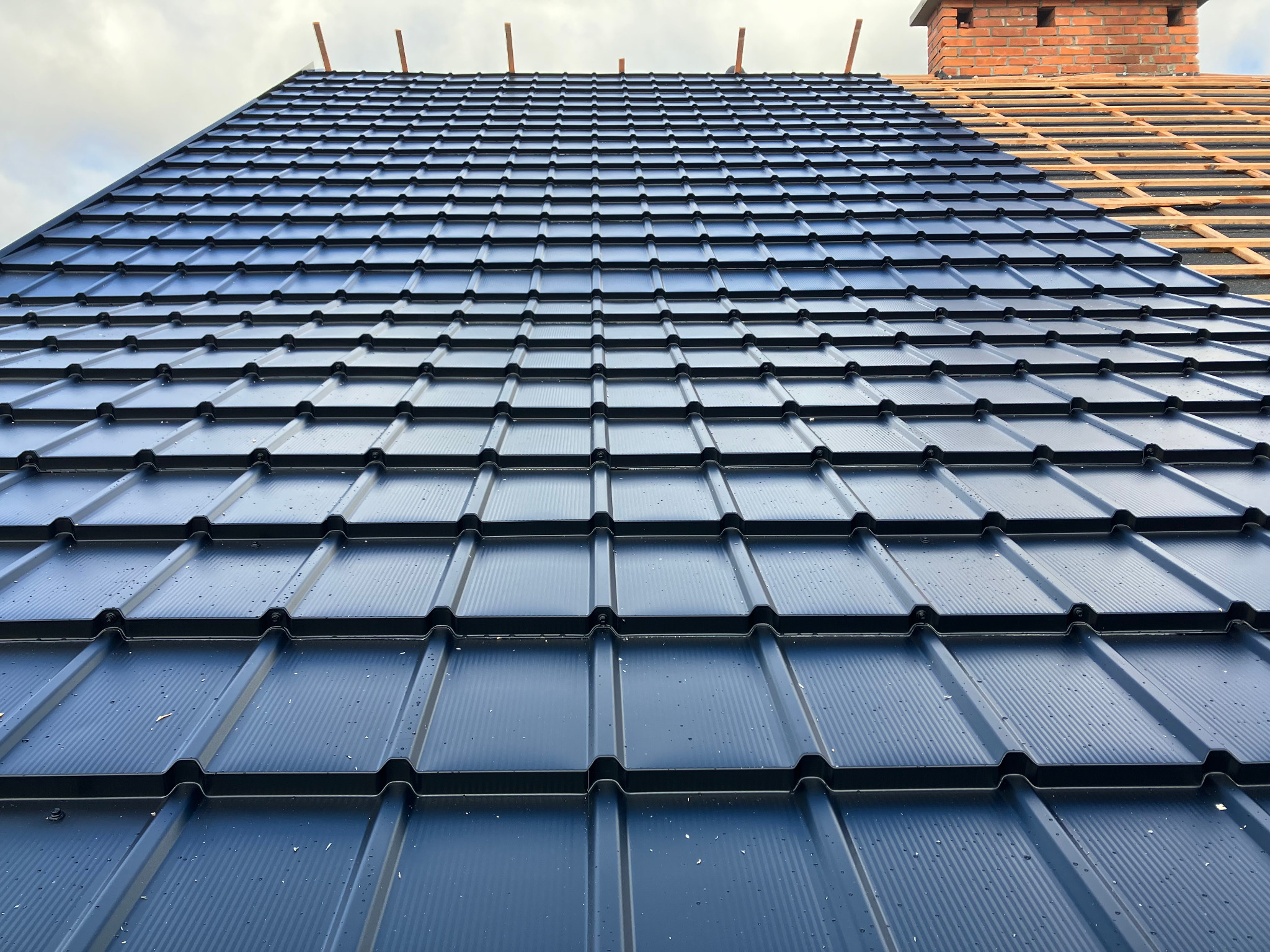 Częściowo pokryty, ciemny dach z blachy trapezowej, widoczny komin z cegły i fragment konstrukcji drewnianej. Ujęcie z dołu, podkreślające regularność wzoru.