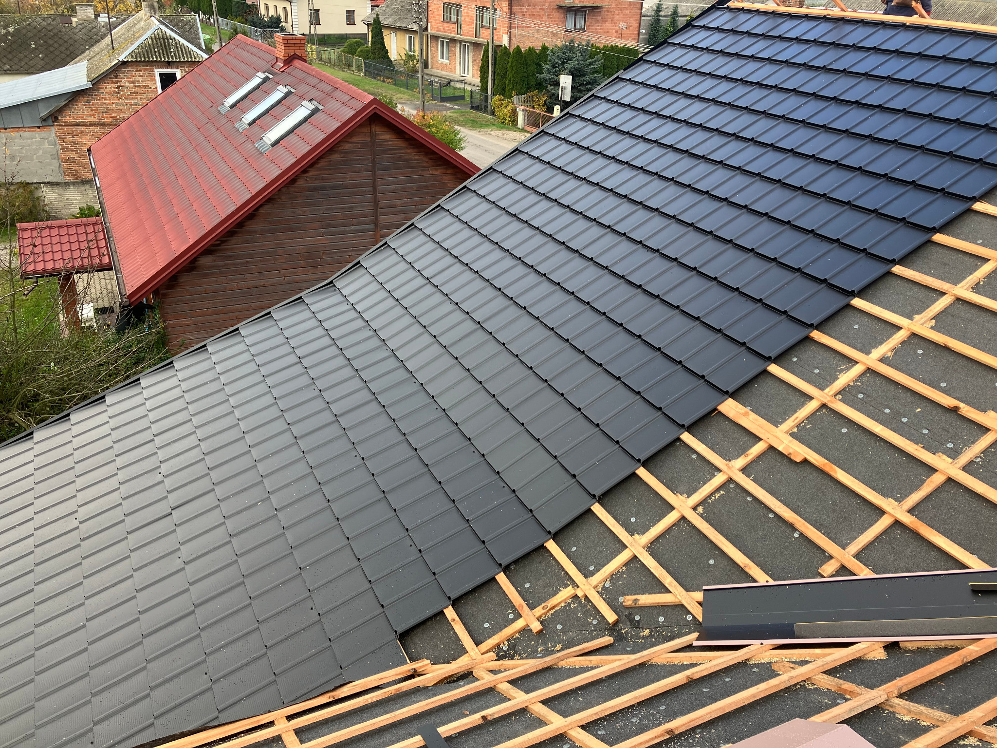 Częściowo pokryty dach blachodachówką w kolorze grafitowym, widoczne łaty i membrana dachowa. W tle budynki mieszkalne i dach z czerwonej dachówki.