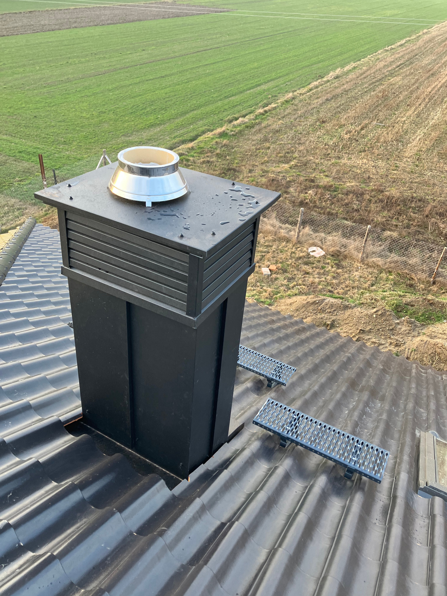 Czarny komin z metalowym zakończeniem na błyszczącym, ciemnym dachu z dachówkami. W tle pole i ogrodzenie. Widoczne stopnie kominiarskie.