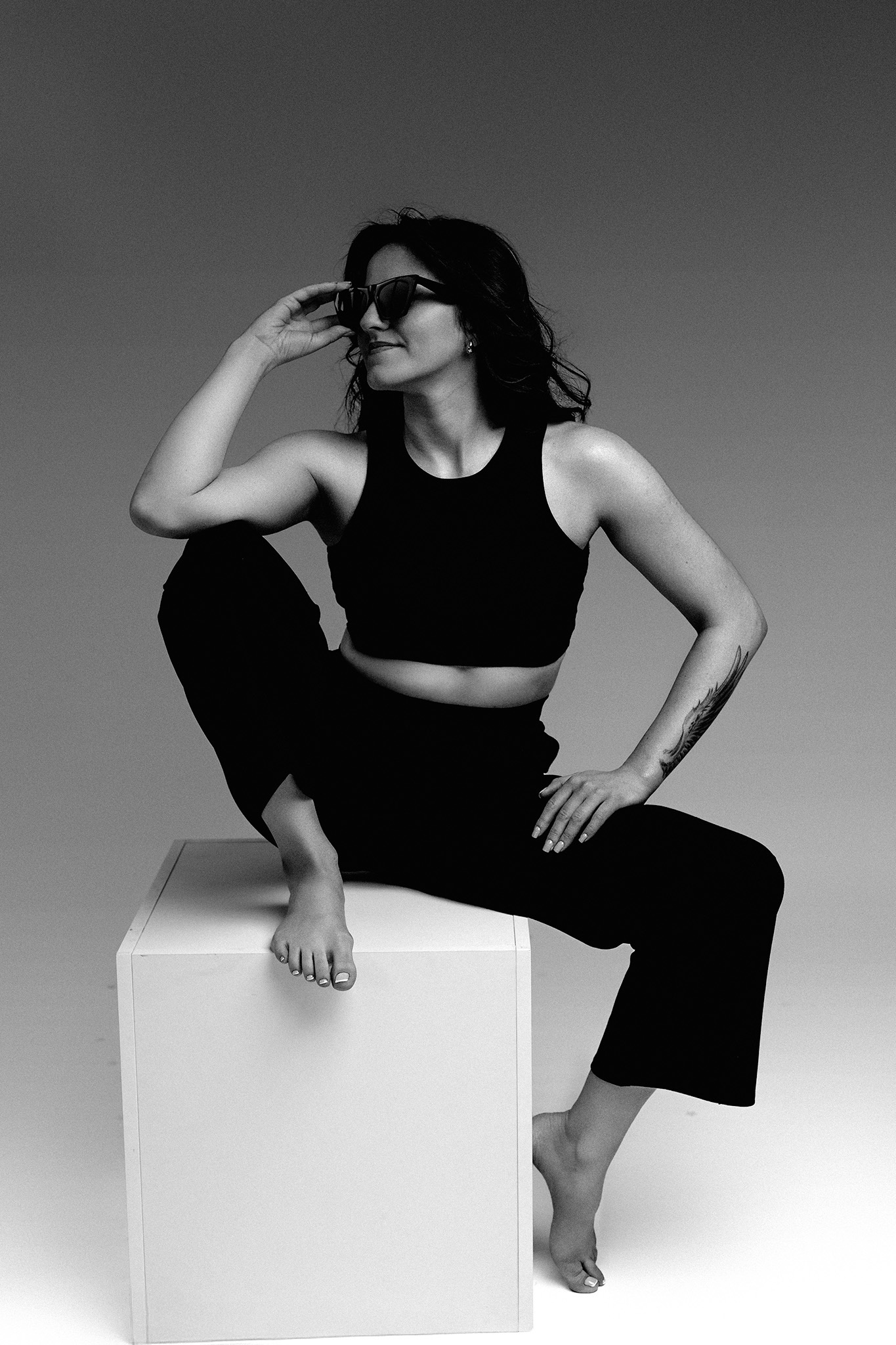Czarno-białe zdjęcie kobiety w okularach przeciwsłonecznych, siedzącej na białym sześcianie, z tatuażem na ramieniu i boso, spoglądającej w bok z uśmiechem.