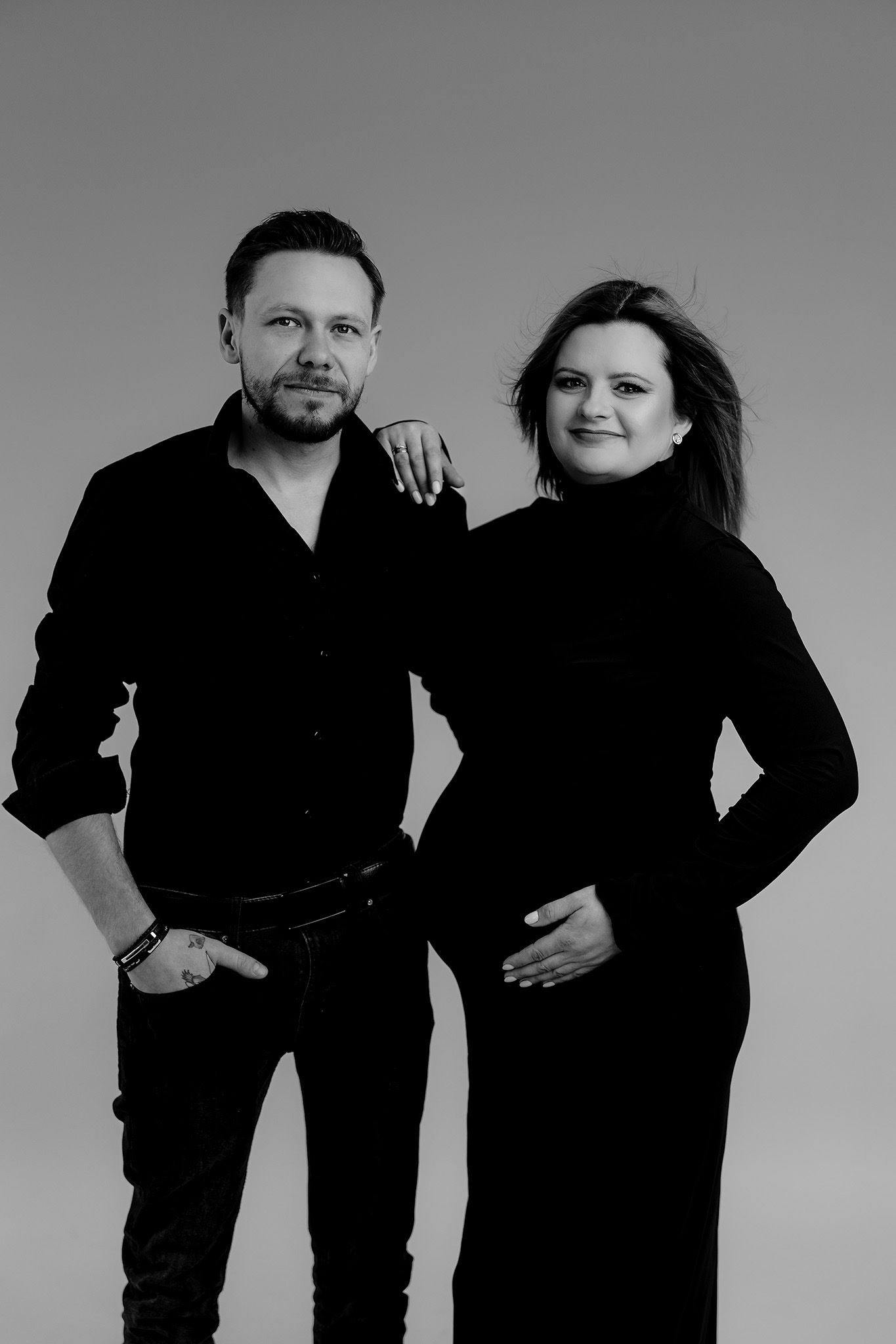 Czarno-biała sesja ciążowa: przyszła mama w czarnej sukience z golfem, dłoń na brzuchu, obok partner z ręką w kieszeni jeansów, subtelny uśmiech, minimalistyczne tło.