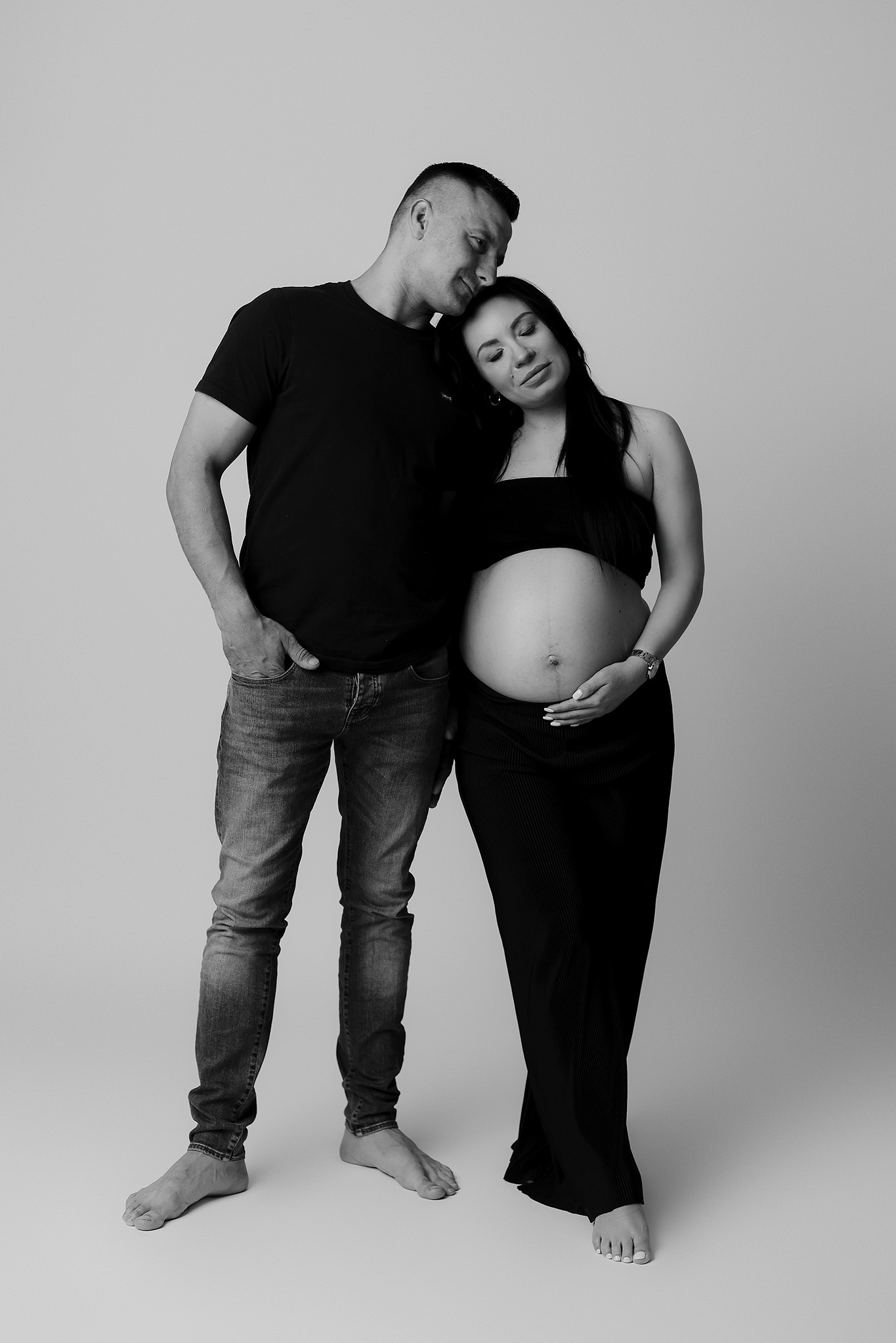 Czarno-białe zdjęcie pary: mężczyzna w jeansach i kobieta w ciąży w czarnej sukience, pozują na jasnym tle. Mężczyzna opiera głowę o głowę kobiety, dłoń na brzuchu.