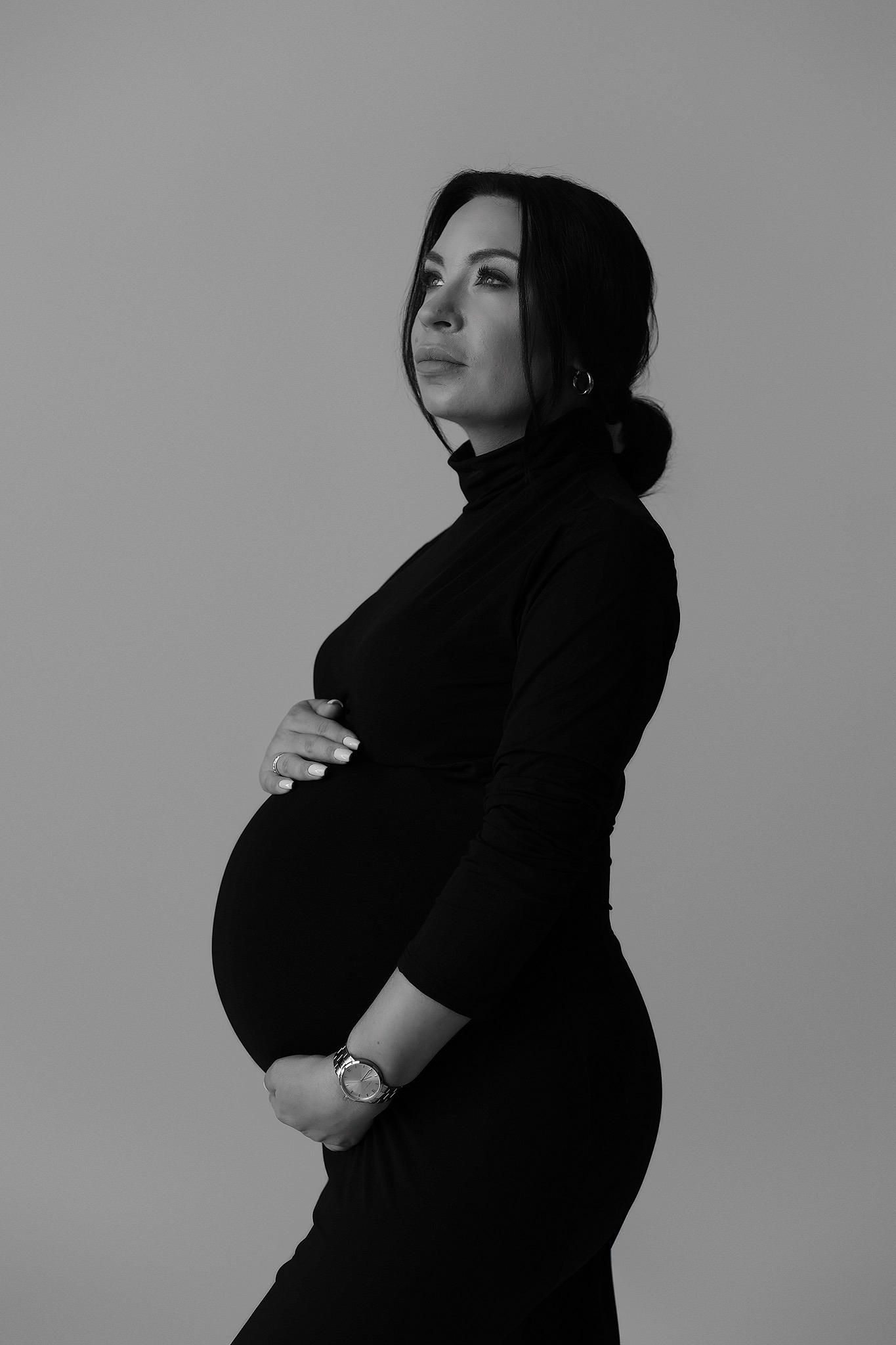 Monochromatyczne, studyjne ujęcie profilowe kobiety w ciąży, ubranej w czarny golf i sukienkę, z dłońmi na brzuchu i zegarkiem na nadgarstku, spoglądającej w górę.