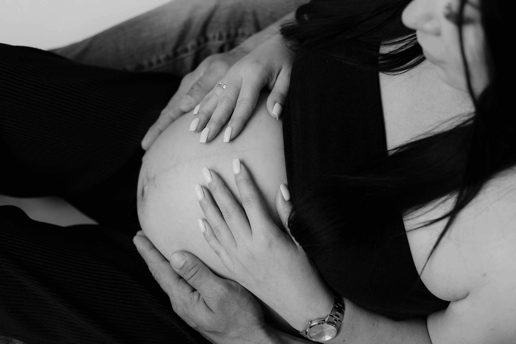 Czarno-białe zbliżenie na ciążowy brzuszek, dłonie kobiety z pierścionkiem i mężczyzny delikatnie go obejmują. Widoczny zegarek na nadgarstku mężczyzny.