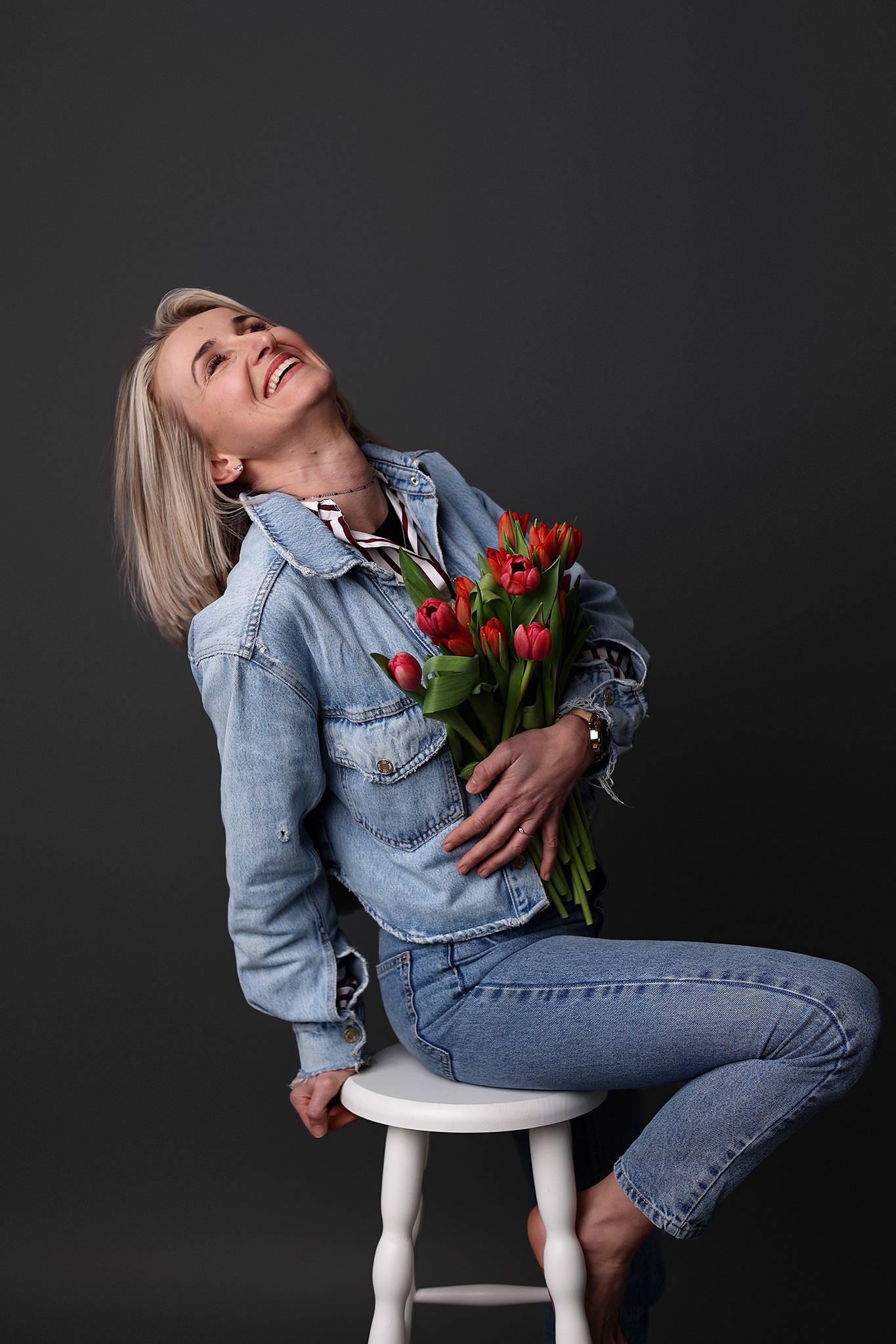 Uśmiechnięta blondynka w jeansowej kurtce, trzymająca bukiet czerwonych tulipanów, siedzi na białym stołku w studio w Mińsku Mazowieckim. Sesja kobieca.