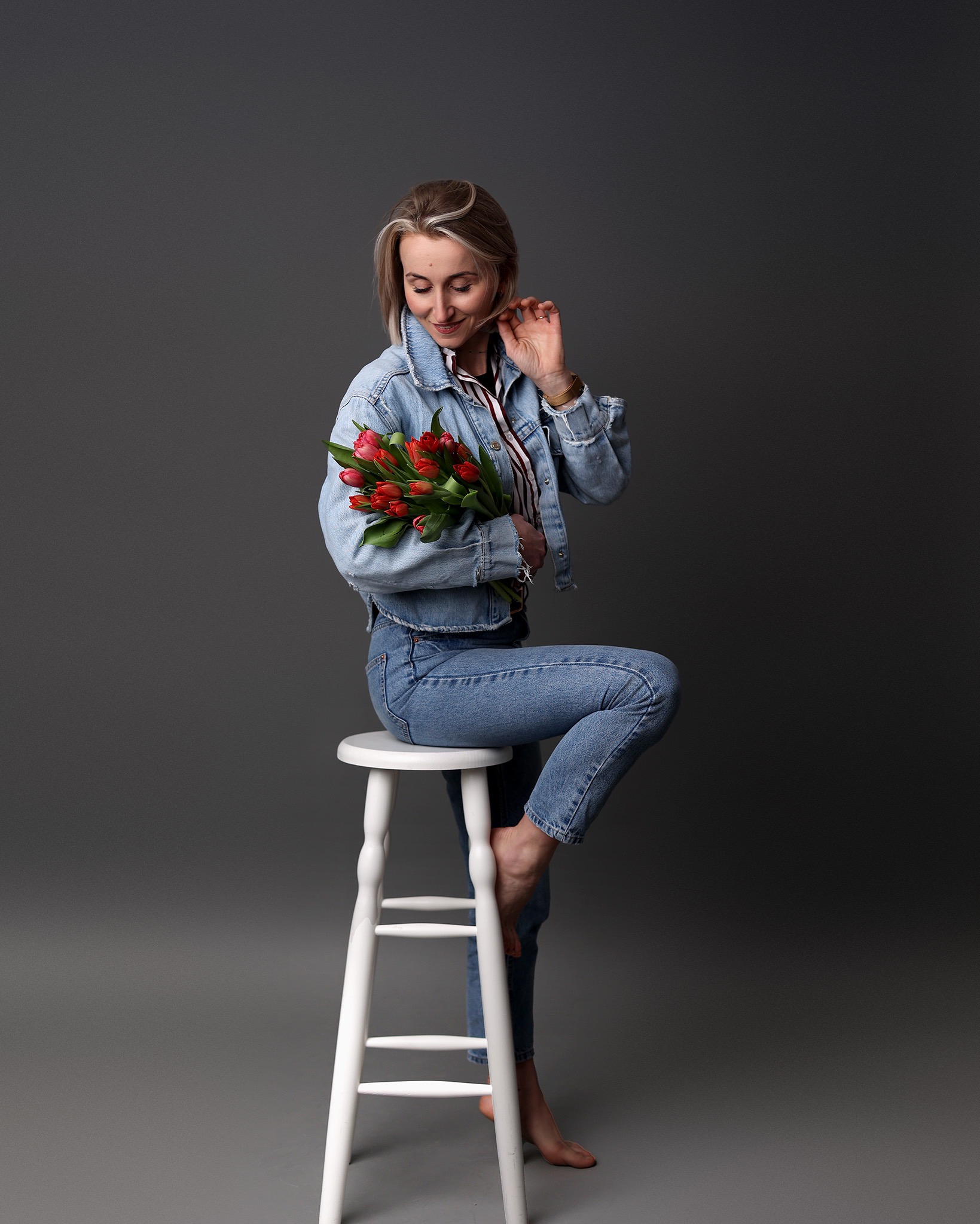 Kobieta w jeansowej kurtce siedzi na białym stołku, trzymając bukiet czerwonych tulipanów na szarym tle. Uśmiechnięta, patrzy na kwiaty, dotykając włosów.