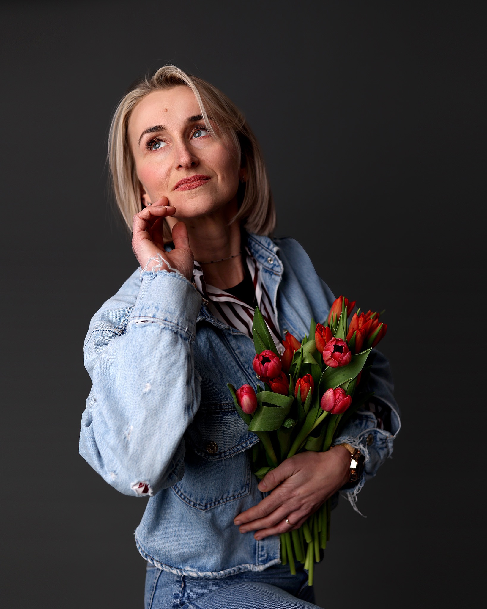 Portret kobiety w jeansowej kurtce, trzymającej bukiet czerwonych tulipanów, zamyślona, spogląda w górę na szarym tle. Widoczny zegarek i pierścionek.