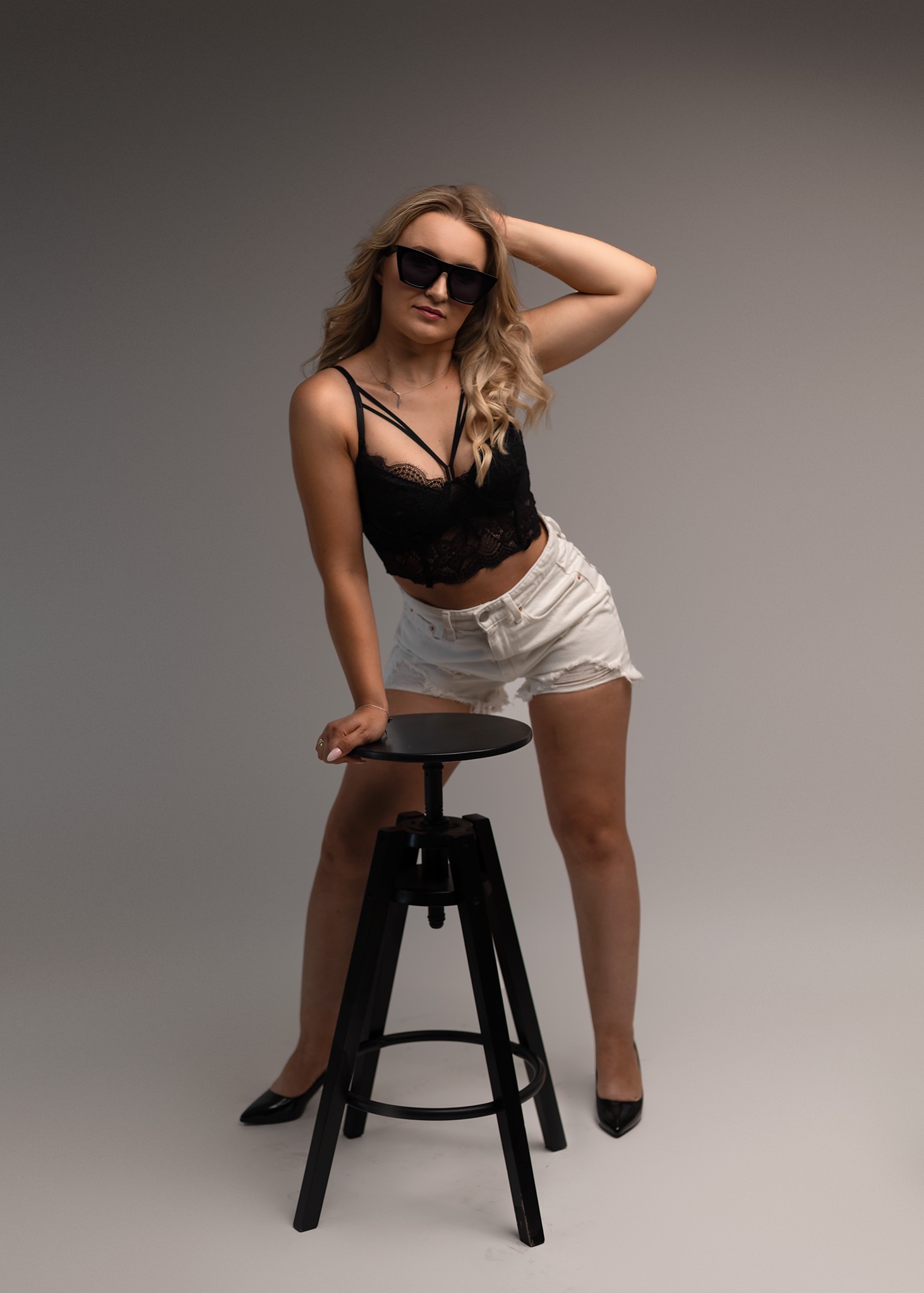 Blondynka w czarnej koronkowej bluzce i białych szortach pozuje opierając się o czarny stołek barowy, w okularach przeciwsłonecznych na szarym tle.