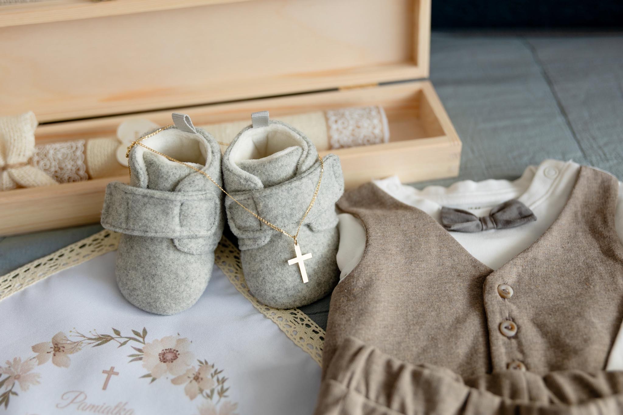 Ubranko do chrztu: szare buciki z krzyżykiem, beżowa kamizelka z muszką, świeca i pamiątkowa serwetka w drewnianym pudełku. Delikatne kolory, bliski kadr.