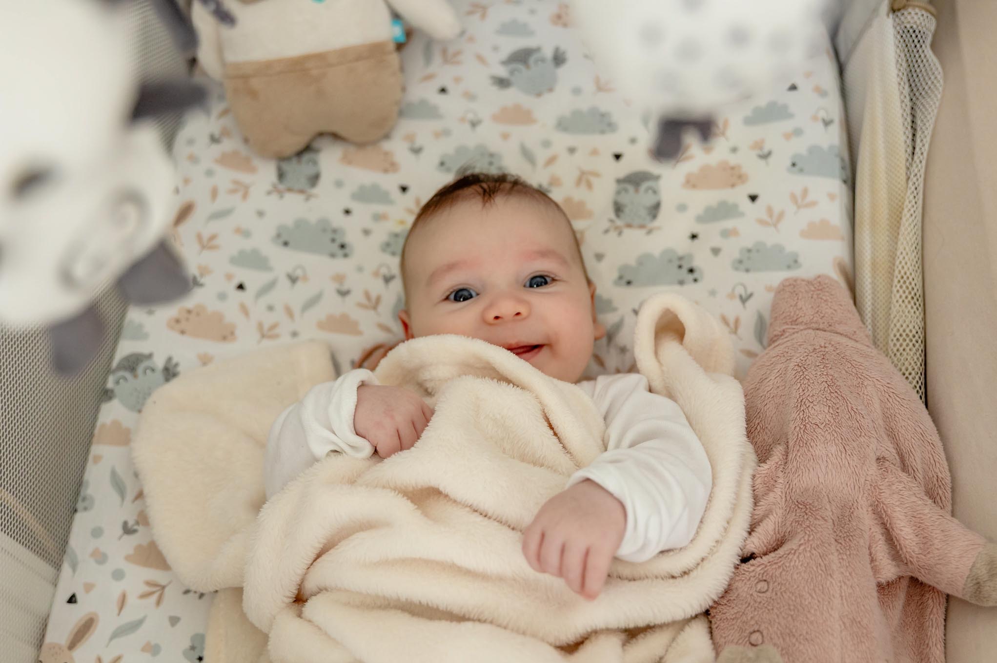Uśmiechnięty niemowlak w łóżeczku otoczony pluszakami, sfotografowany z góry w Mińsku Mazowieckim. Dziecko przykryte miękkim, kremowym kocykiem.
