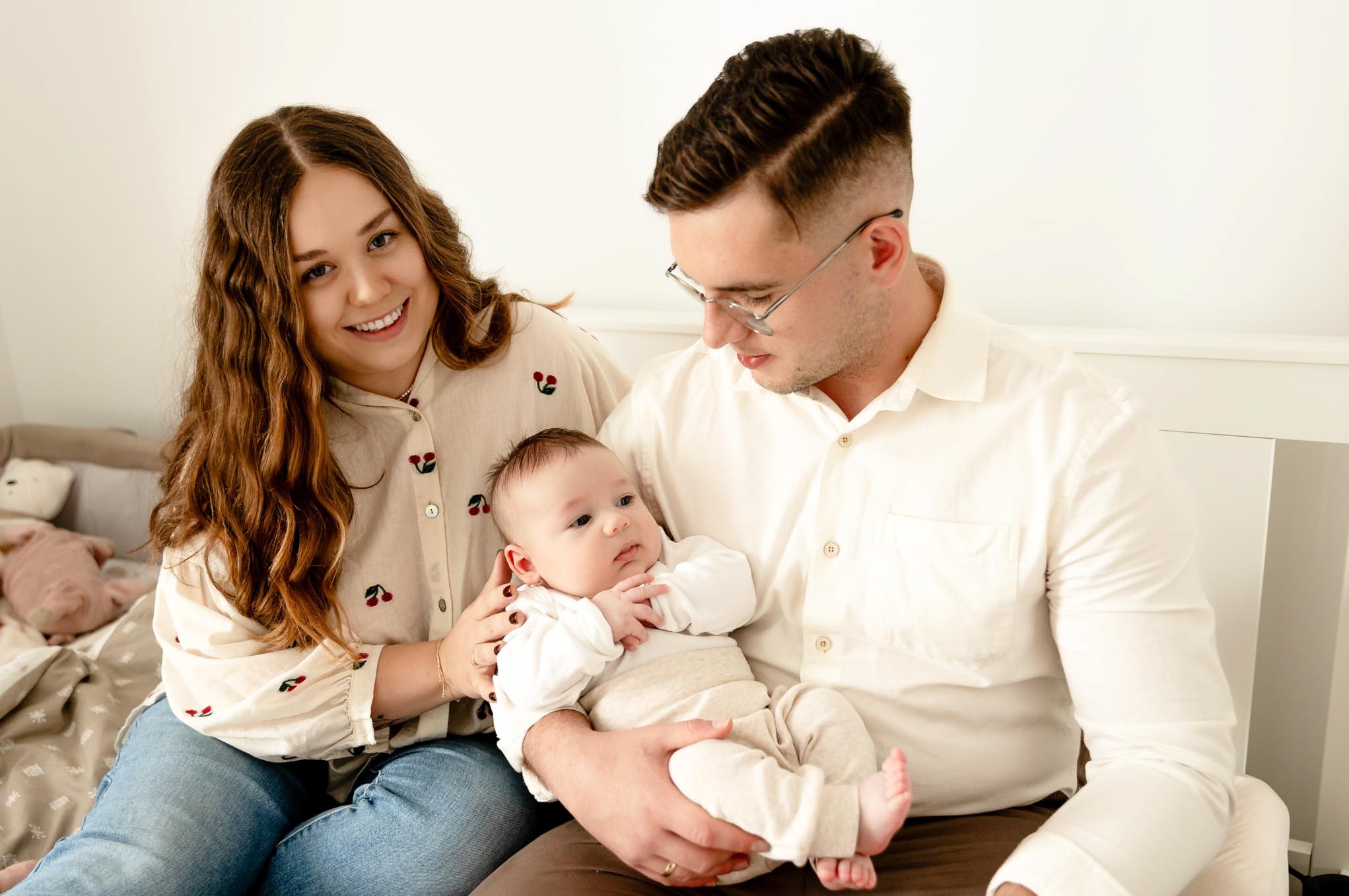Uśmiechnięta mama, tata w okularach trzymający niemowlę w beżowym ubranku, sesja rodzinna w jasnym wnętrzu, naturalne światło.