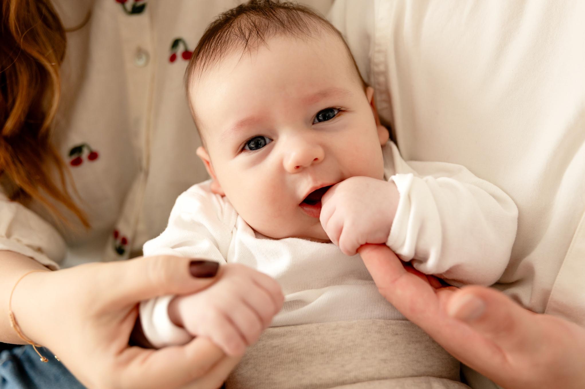 Urocze niemowlę w ramionach mamy, z palcem w buzi, w ciepłym oświetleniu. Ubrana w bluzkę w wiśnie, delikatny uśmiech dziecka.