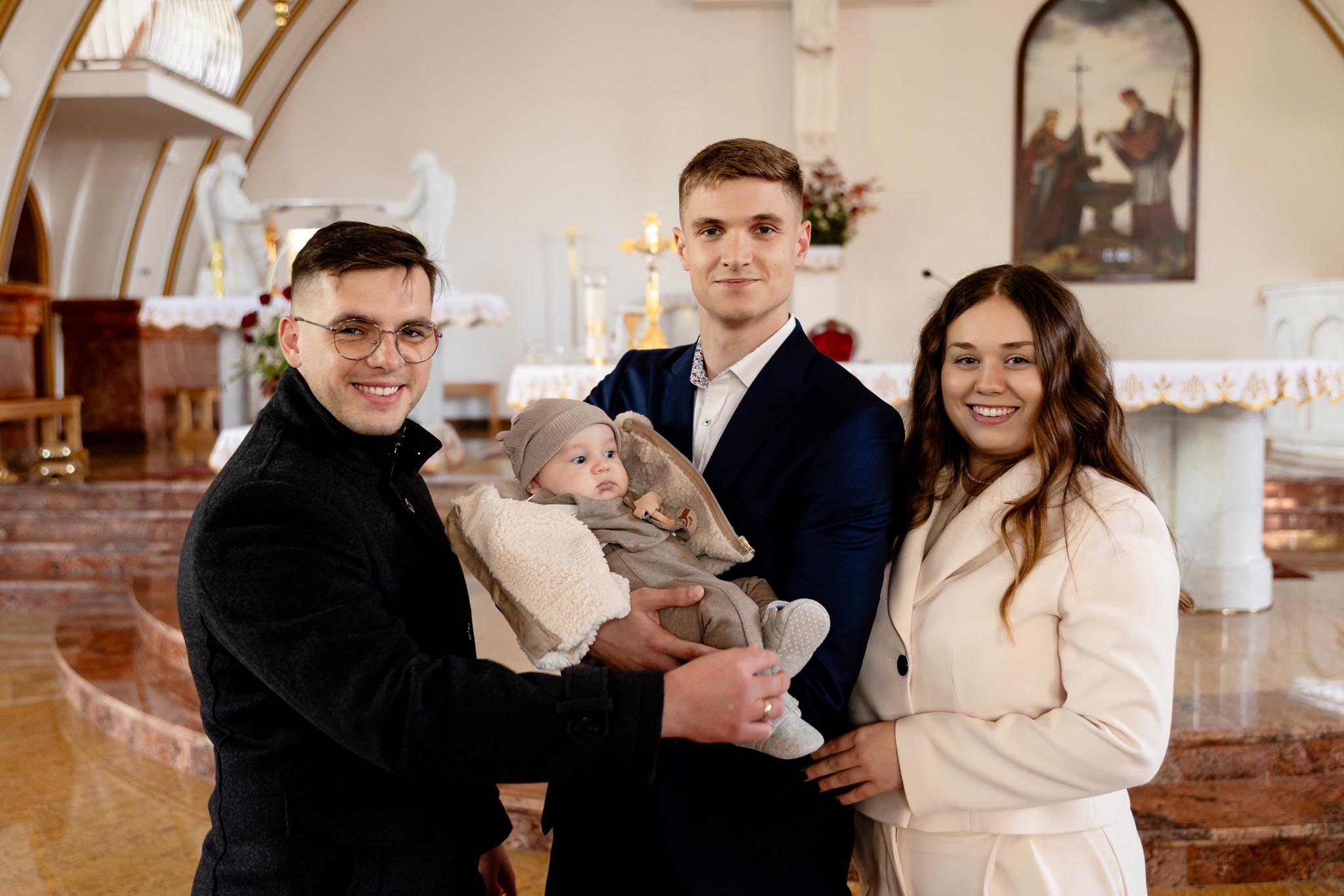 Uśmiechnięta rodzina z niemowlęciem w kościele, prawdopodobnie po ceremonii chrztu. Ojciec trzyma dziecko, obok stoją rodzice chrzestni. W tle ołtarz i dekoracje.