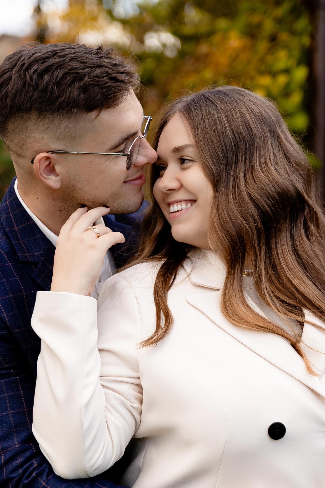 Uśmiechnięta para w zbliżeniu: ona w kremowej marynarce, on w okularach i granatowej marynarce w kratę, delikatnie dotyka jej twarzy. Romantyczna sesja w plenerze.