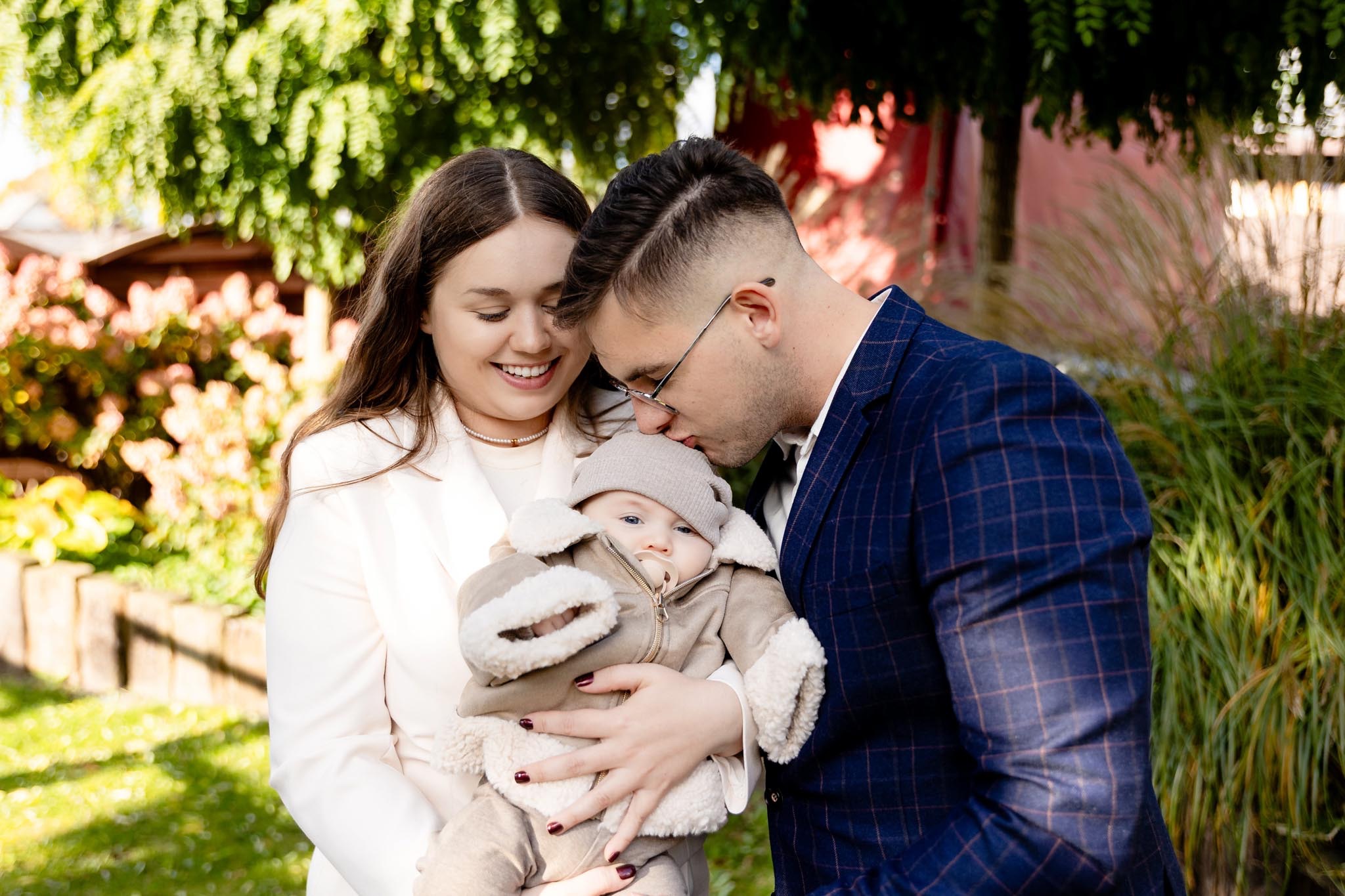 Uśmiechnięta mama w białym żakiecie i tata w kraciastej marynarce trzymają na rękach niemowlę w beżowym kombinezonie na tle zieleni i czerwonego budynku.