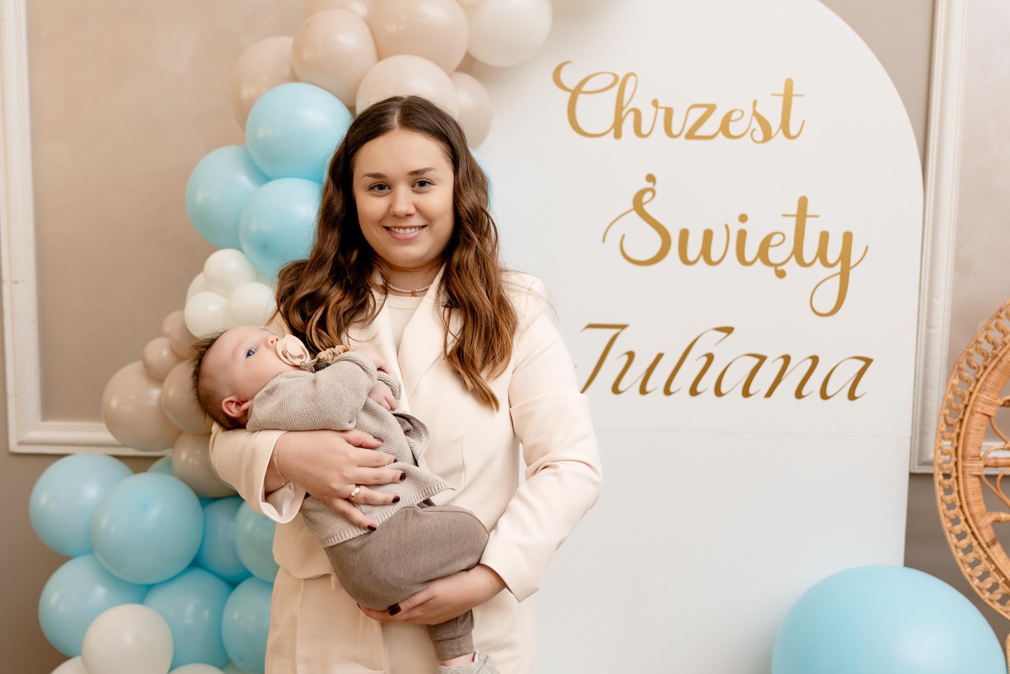 Uśmiechnięta kobieta w kremowym żakiecie trzyma niemowlę na tle dekoracji z balonów i napisu 'Chrzest Święty Juliana' w kolorze złota.