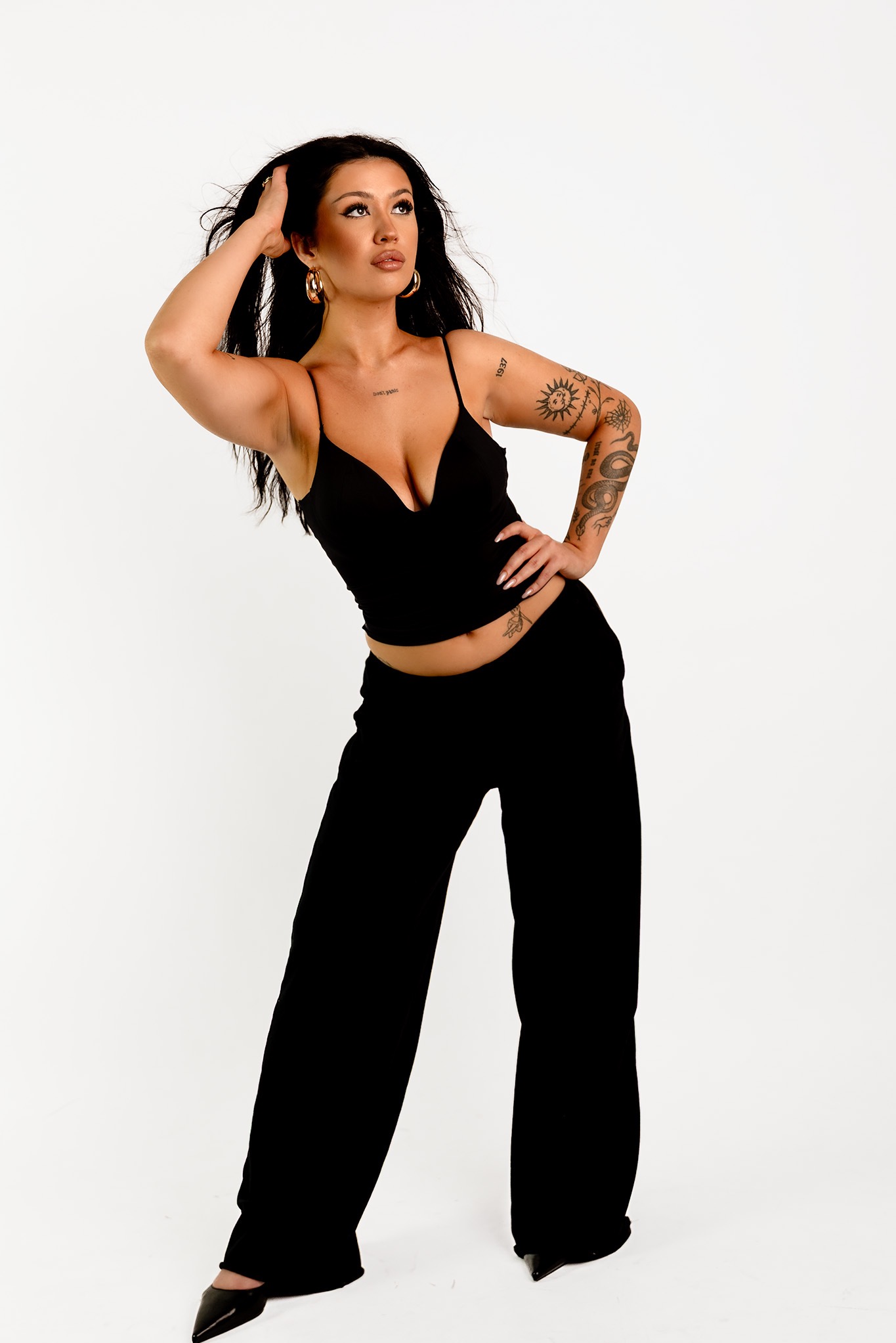 Kobieta w czarnym stroju pozuje na białym tle, z ręką we włosach i tatuażami na ramieniu. Widoczne złote kolczyki i delikatny makijaż. Ujęcie z dołu.