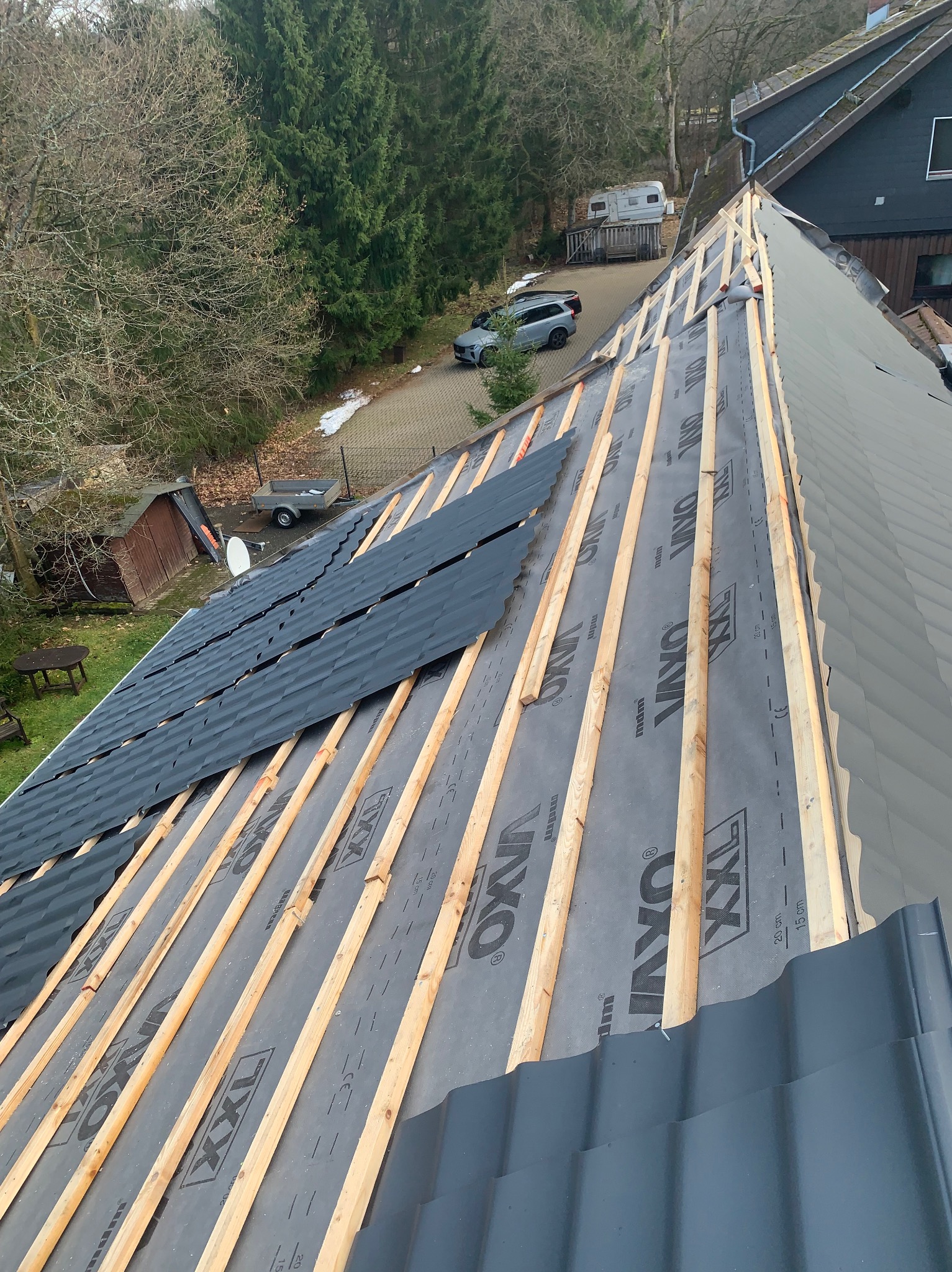 Montaż pokrycia dachowego z czarnej blachy na łatach i membranie. Widok z góry na częściowo ukończony dach z widocznymi elementami konstrukcyjnymi.