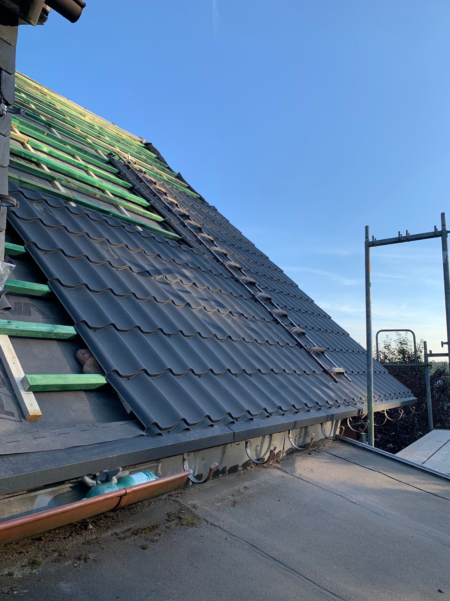 Częściowo pokryty dach z czarnej blachy falistej, widoczne zielone łaty, rusztowanie i rynna. Ujęcie z dołu, błękitne niebo w tle.