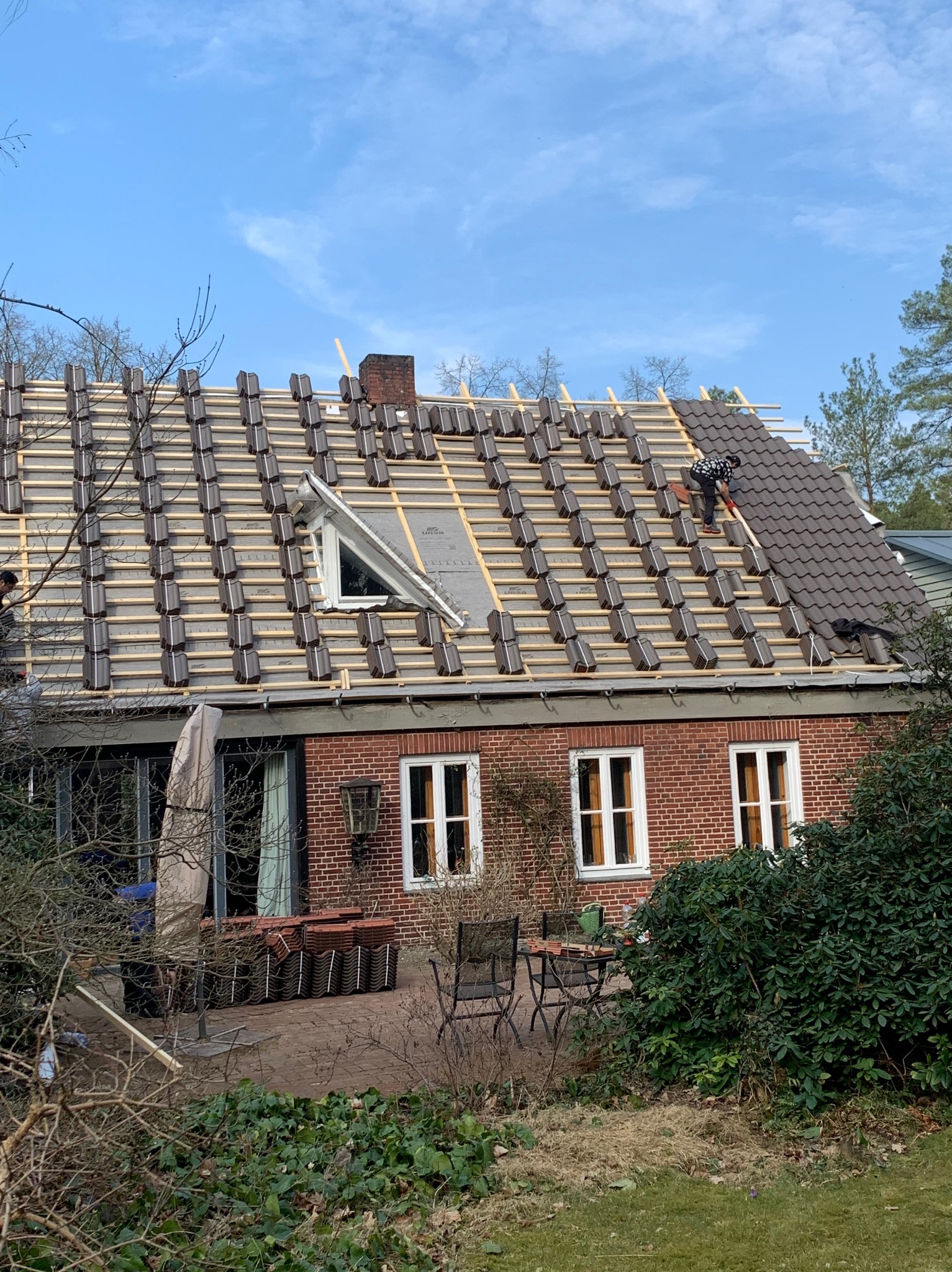 Układanie brązowej dachówki na drewnianej konstrukcji dachu domu z czerwonej cegły. Część dachu pokryta, część w trakcie. Widoczne okno dachowe i komin.