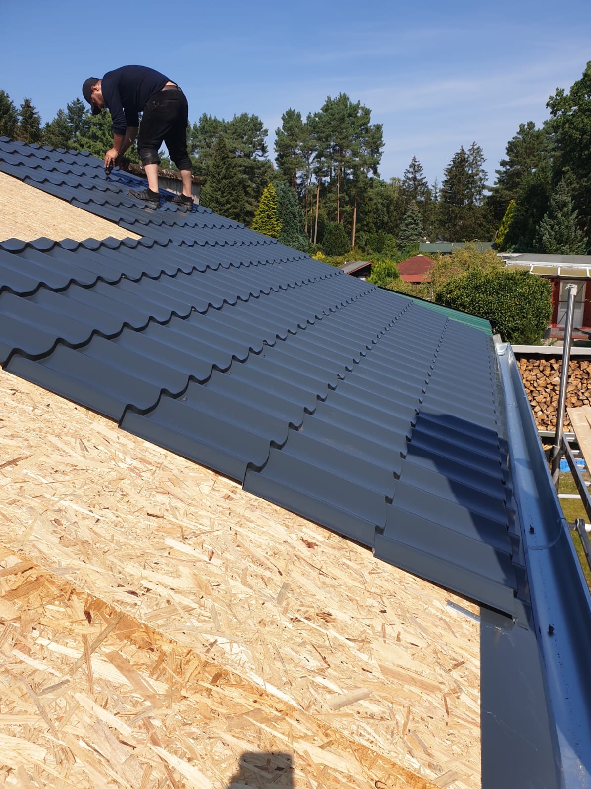 Dekarz montuje ciemnoszarą blachodachówkę na drewnianej konstrukcji dachu. Widoczna rynna i drzewa w tle, błękitne niebo. Praca w toku, etap krycia dachu.