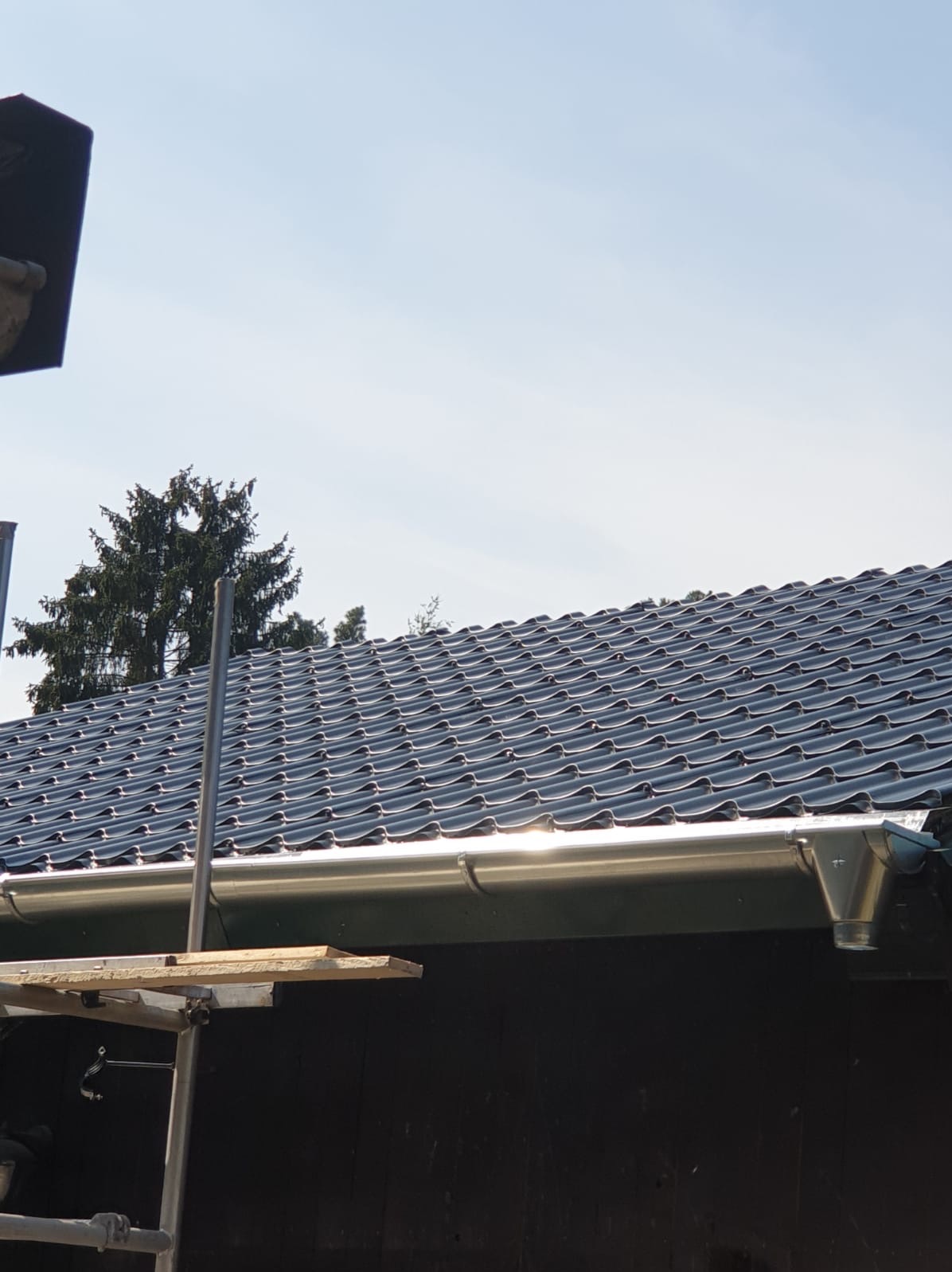 Błyszczące, grafitowe dachówki ceramiczne na skośnym dachu z zamontowaną, metalową rynną. Fragment rusztowania w lewym dolnym rogu. Błękitne niebo w tle.