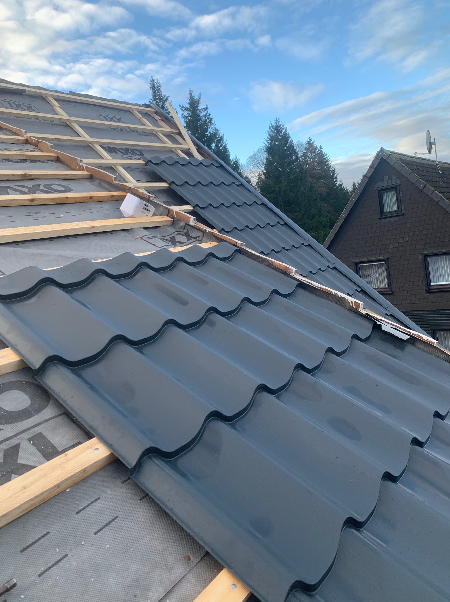 Montaż ciemnoszarej blachodachówki na konstrukcji drewnianej z membraną, widok z góry, w tle dom i drzewa. Częściowo pokryty dach w trakcie prac dekarskich.