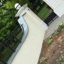 Jakub Taczka - Eleganckie ogrodzenie z czarną, kutą bramą i oświetlonym słupkiem, obok ścieżka z kostki brukowej i rabata z roślinami. Jasna elewacja budynku w tle.