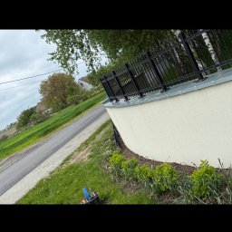 Jakub Taczka - Nowoczesne ogrodzenie z czarnymi metalowymi przęsłami, zamontowane na niskim, białym murku okalającym posesję. Widoczne narzędzia w wiadrze obok.