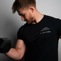 Maciej Maśko - Mężczyzna w czarnej koszulce z napisem 'Trener Personalny Maciej Maśko' ćwiczy biceps z hantlą na tle szarej ściany. Widoczny napięty biceps i skupiony wzrok.