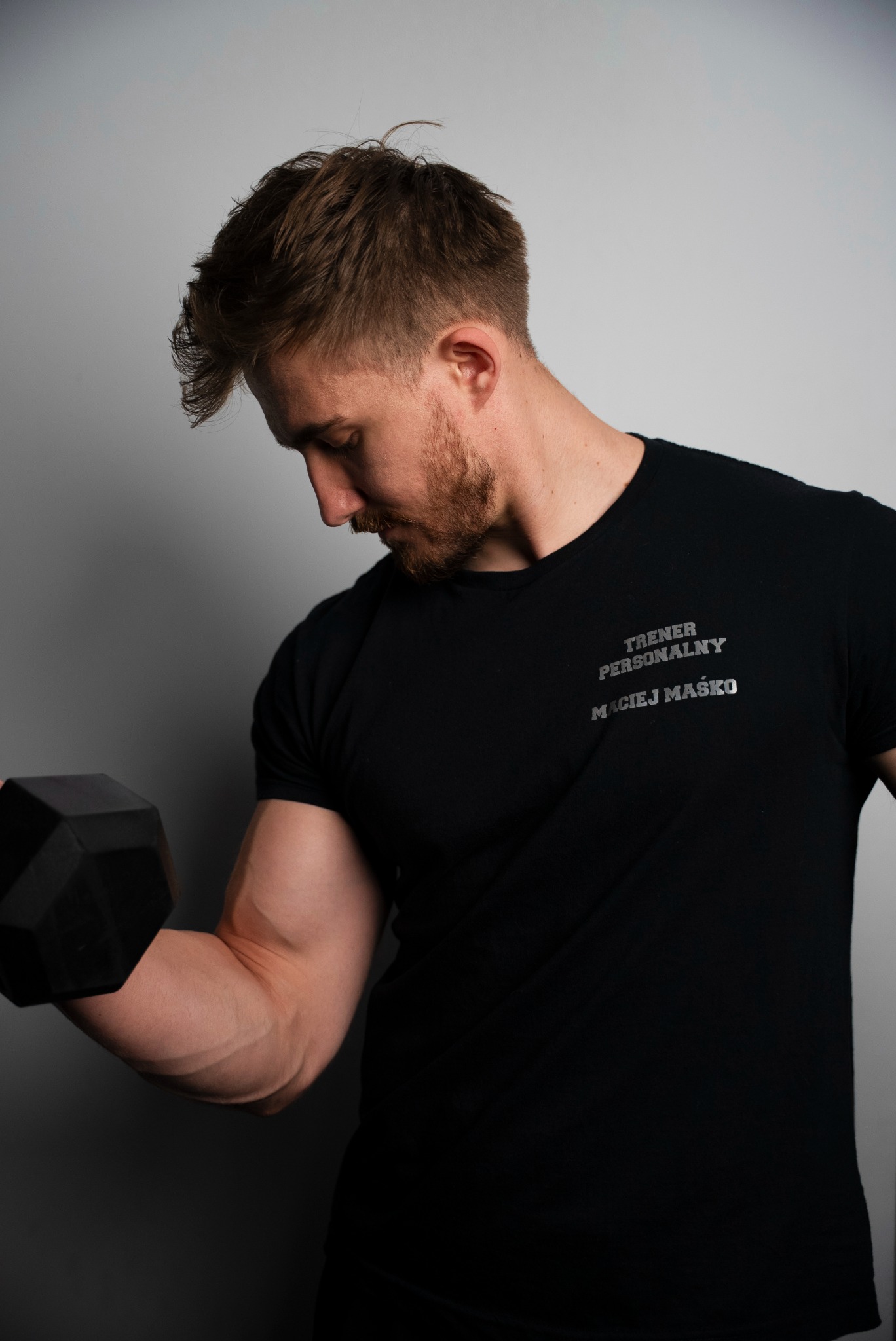 Mężczyzna w czarnej koszulce z napisem 'Trener Personalny Maciej Maśko' ćwiczy biceps z hantlą na tle szarej ściany. Widoczny napięty biceps i skupiony wzrok.