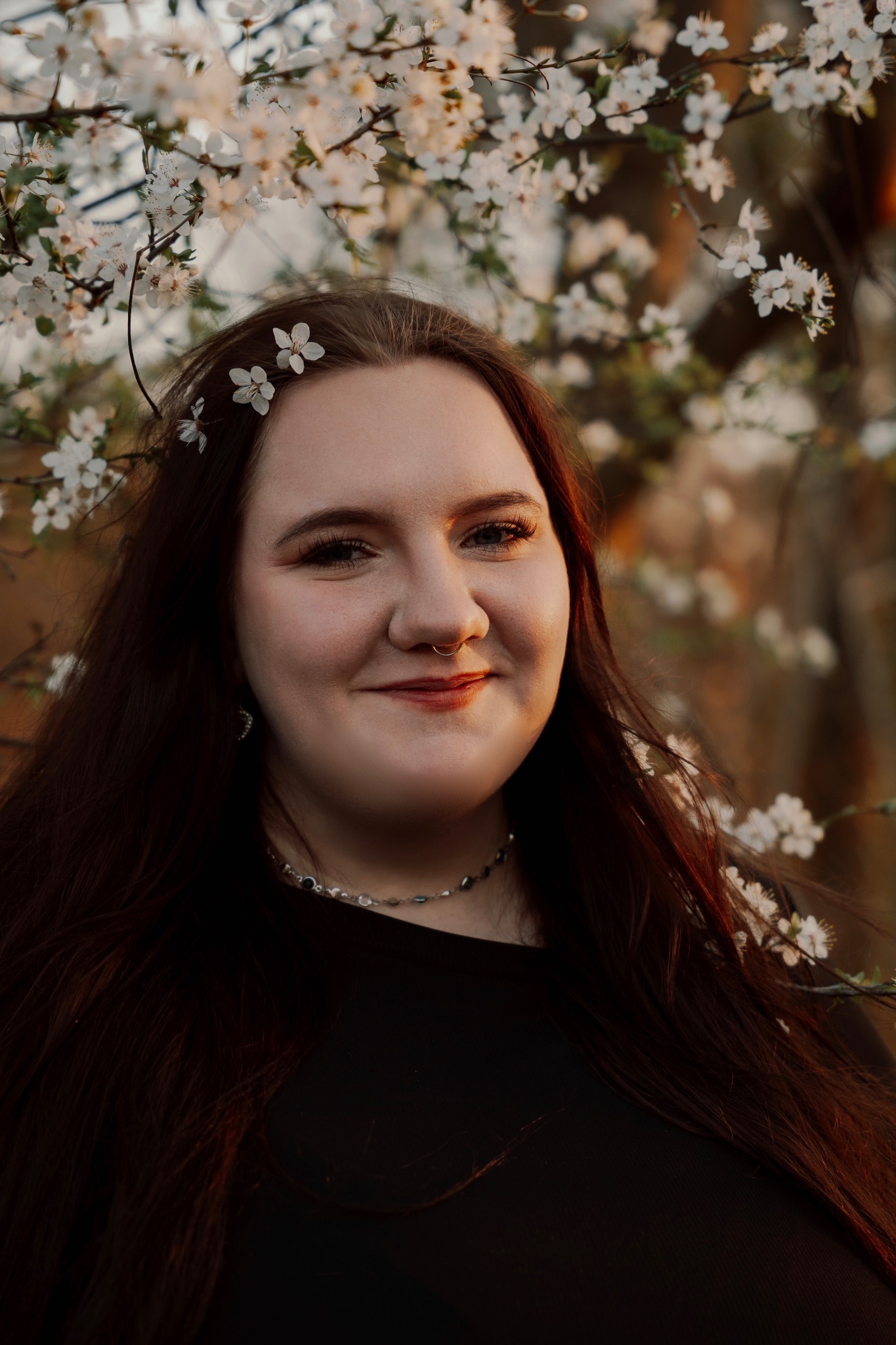 Portret uśmiechniętej kobiety z kwiatami we włosach na tle kwitnącego drzewa w Szczecinie. Naturalne światło, ciepłe barwy, widoczny kolczyk w nosie.