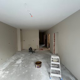 Zav-Bud - Wnętrze po malowaniu ścian na neutralny odcień szarości. Widoczne ślady po remoncie na podłodze, drabina malarska i narzędzia. Otwory na gniazdka elektryczne.