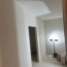 Zav-Bud - Wnętrze po szpachlowaniu ścian i sufitu, przygotowane do malowania. Widoczny reflektor budowlany i ślady po pracach wykończeniowych na podłodze.
