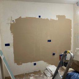 Zav-Bud - Ściana w trakcie szpachlowania z widocznymi niebieskimi punktami mocowania, drabina i materiały budowlane w pomieszczeniu. Prace wykończeniowe w trakcie.