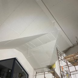 Zav-Bud - Skośny sufit z płyt karton-gips, widoczne łączenia i wkręty, rusztowanie w rogu. Ukończony etap prac w pomieszczeniu ze skośnym oknem.