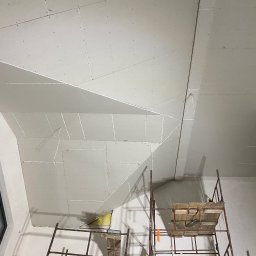 Zav-Bud - Skośny sufit poddasza z zamontowaną płytą kartonowo-gipsową, widoczne łączenia i wkręty, rusztowanie w pomieszczeniu, okno w tle. Prace wykończeniowe.