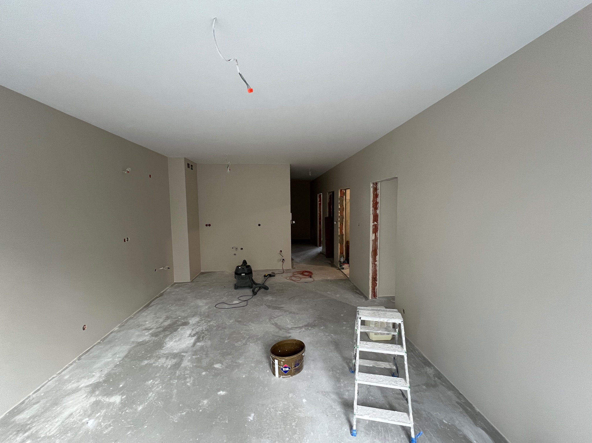 Wnętrze po malowaniu ścian na neutralny odcień szarości. Widoczne ślady po remoncie na podłodze, drabina malarska i narzędzia. Otwory na gniazdka elektryczne.