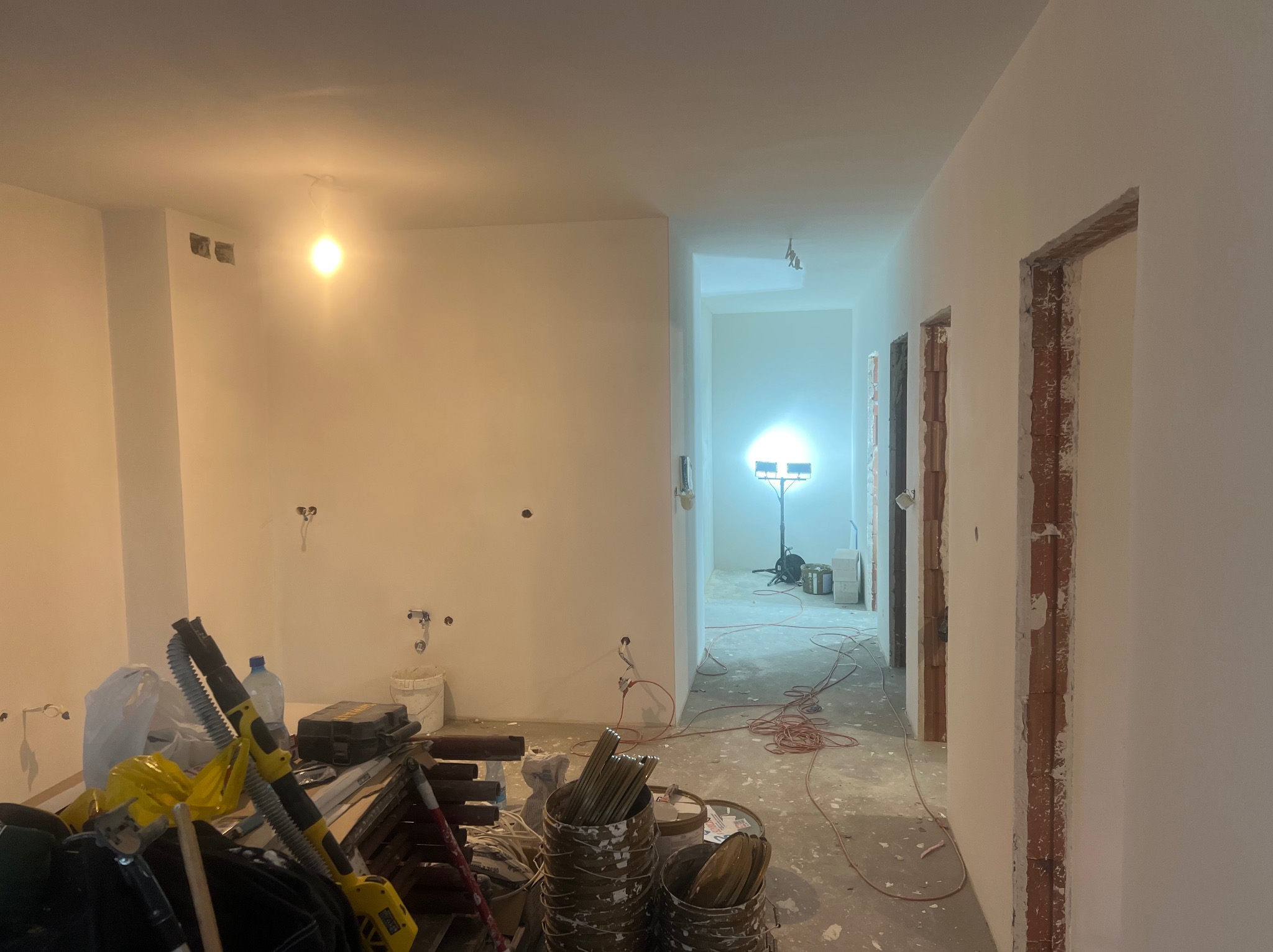 Wnętrze w trakcie malowania ścian na biało. Widoczne narzędzia malarskie, kable i lampa budowlana. Otwory po gniazdkach elektrycznych.