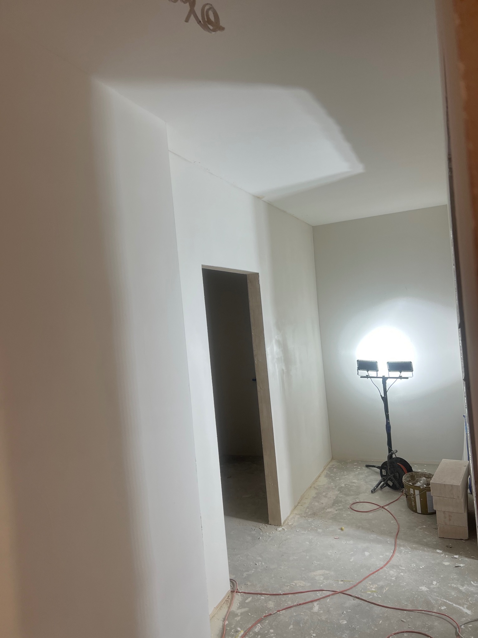 Wnętrze po szpachlowaniu ścian i sufitu, przygotowane do malowania. Widoczny reflektor budowlany i ślady po pracach wykończeniowych na podłodze.