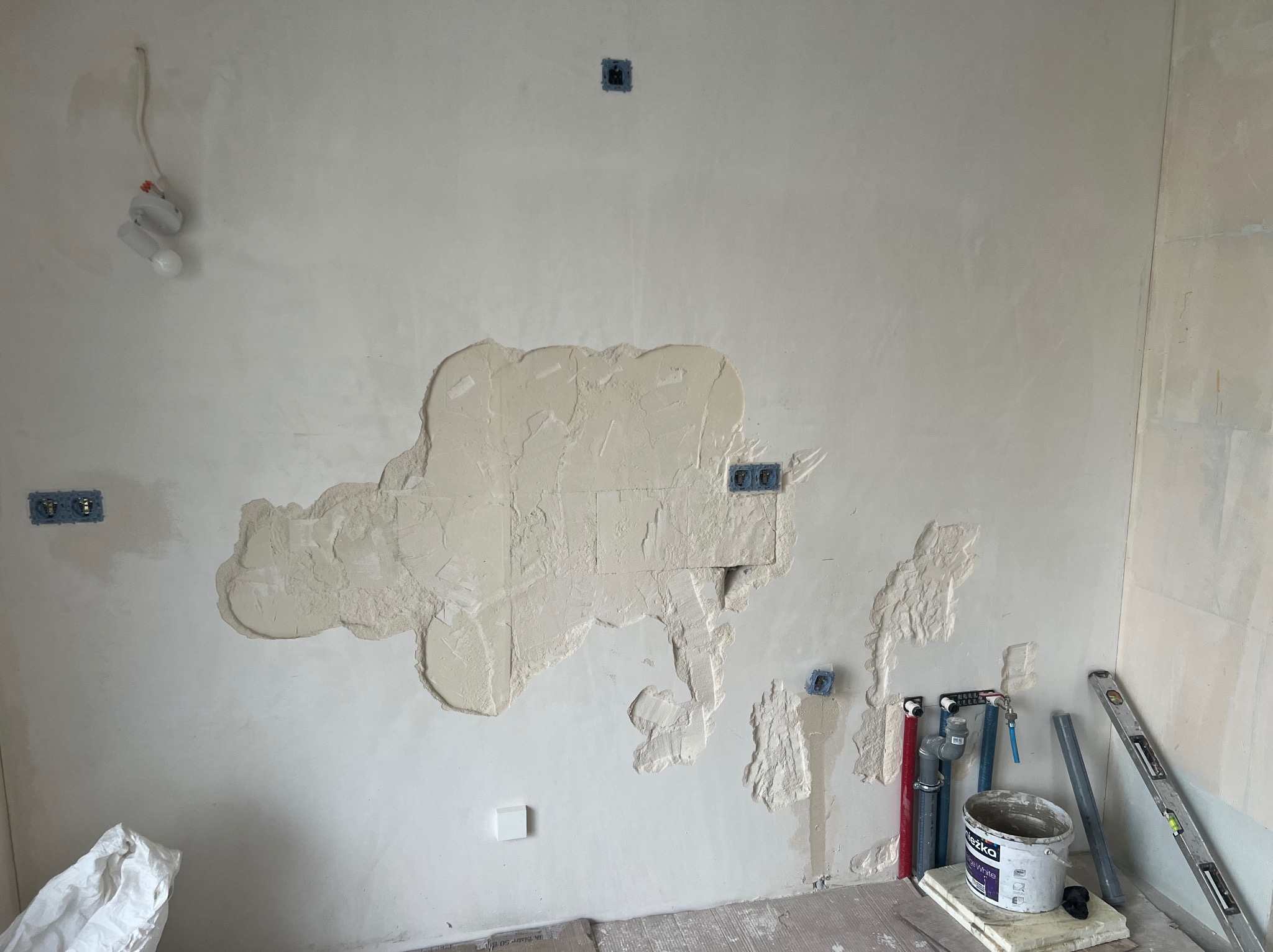 Ściana w trakcie szpachlowania z widocznymi puszkami elektrycznymi, rurami i narzędziami. Wiadro z masą szpachlową stoi na podłodze. Widoczne nierówności i faktura na ścianie.