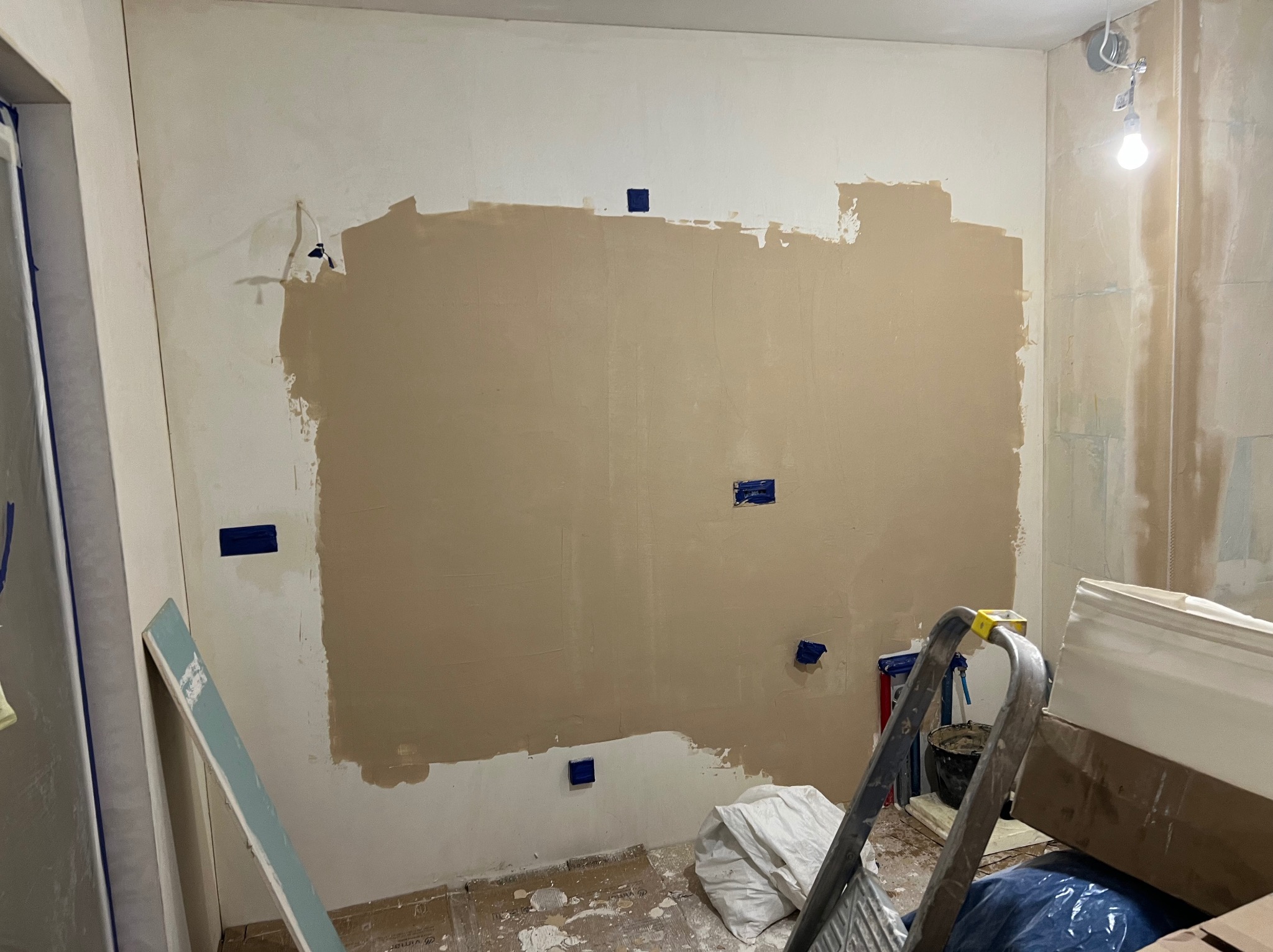 Ściana w trakcie szpachlowania z widocznymi niebieskimi punktami mocowania, drabina i materiały budowlane w pomieszczeniu. Prace wykończeniowe w trakcie.