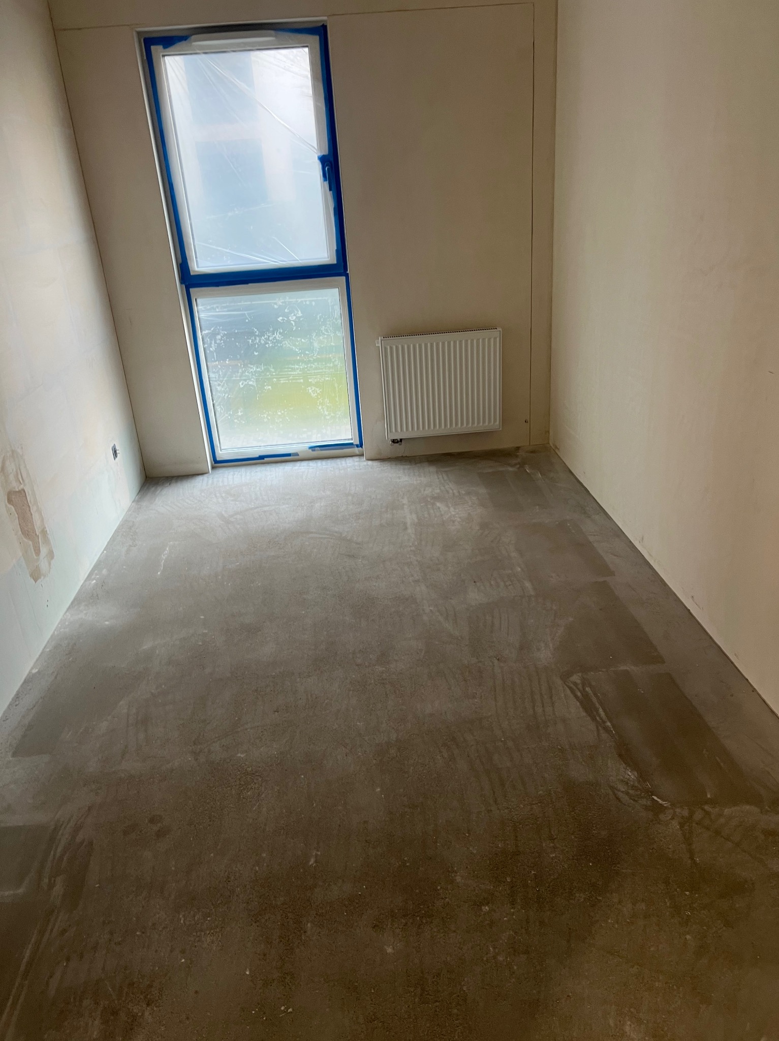 Pokój w trakcie remontu: ściany przygotowane do malowania, okno zabezpieczone folią, grzejnik. Podłoga pokryta wylewką samopoziomującą. Widoczne ślady po pracy.