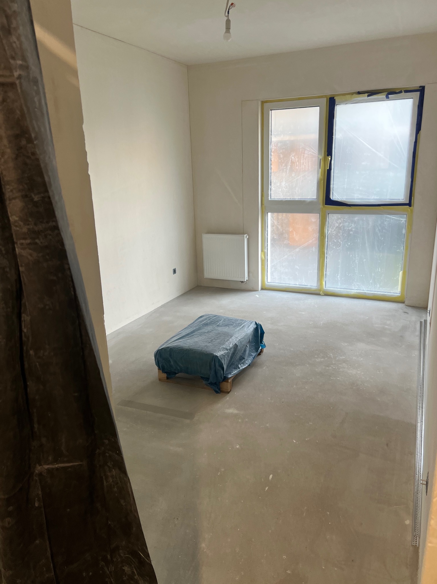Pokój w trakcie prac wykończeniowych: zabezpieczone okno folią i taśmą, podłoga przygotowana pod wylewkę, ściany po szpachlowaniu, grzejnik. Widok zza osłony.