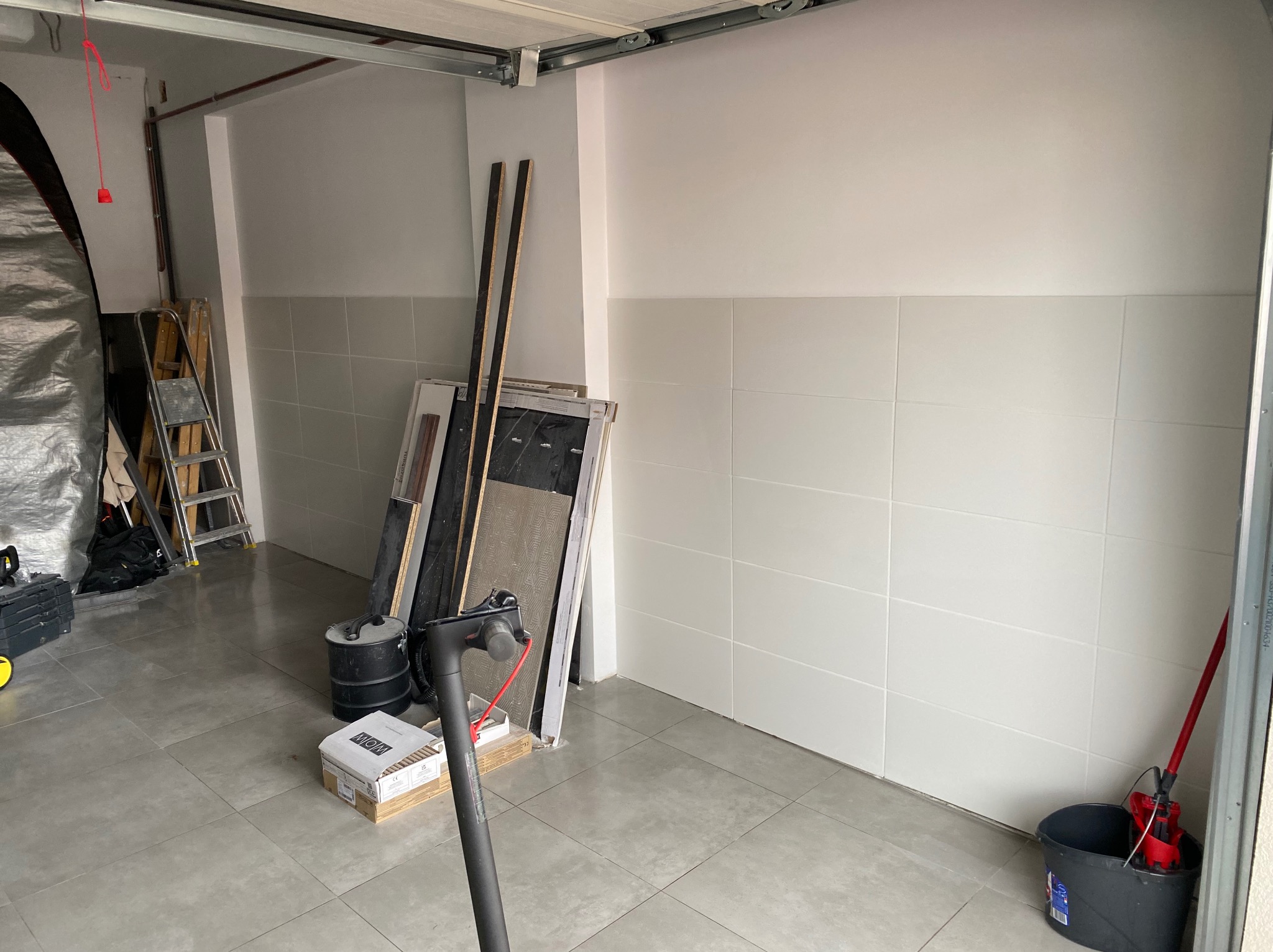 Ściana wyłożona białymi płytkami w garażu, widoczne narzędzia i materiały budowlane, podłoga wyłożona szarymi płytkami. Przygotowania do prac wykończeniowych.