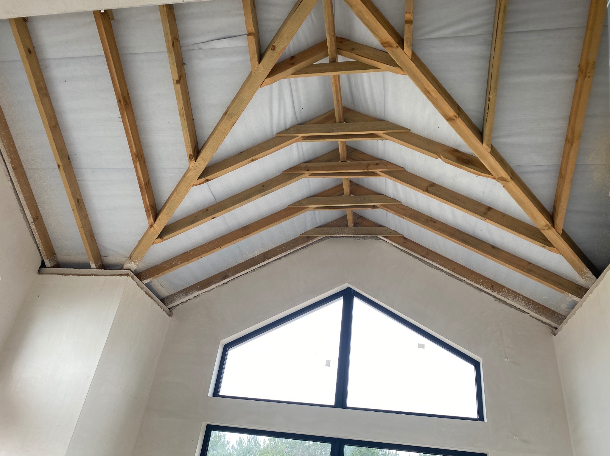 Wnętrze w trakcie prac: surowa ściana z oknem w kształcie trójkąta, widoczna drewniana konstrukcja dachu z membraną. Etap przygotowania do wykończenia.